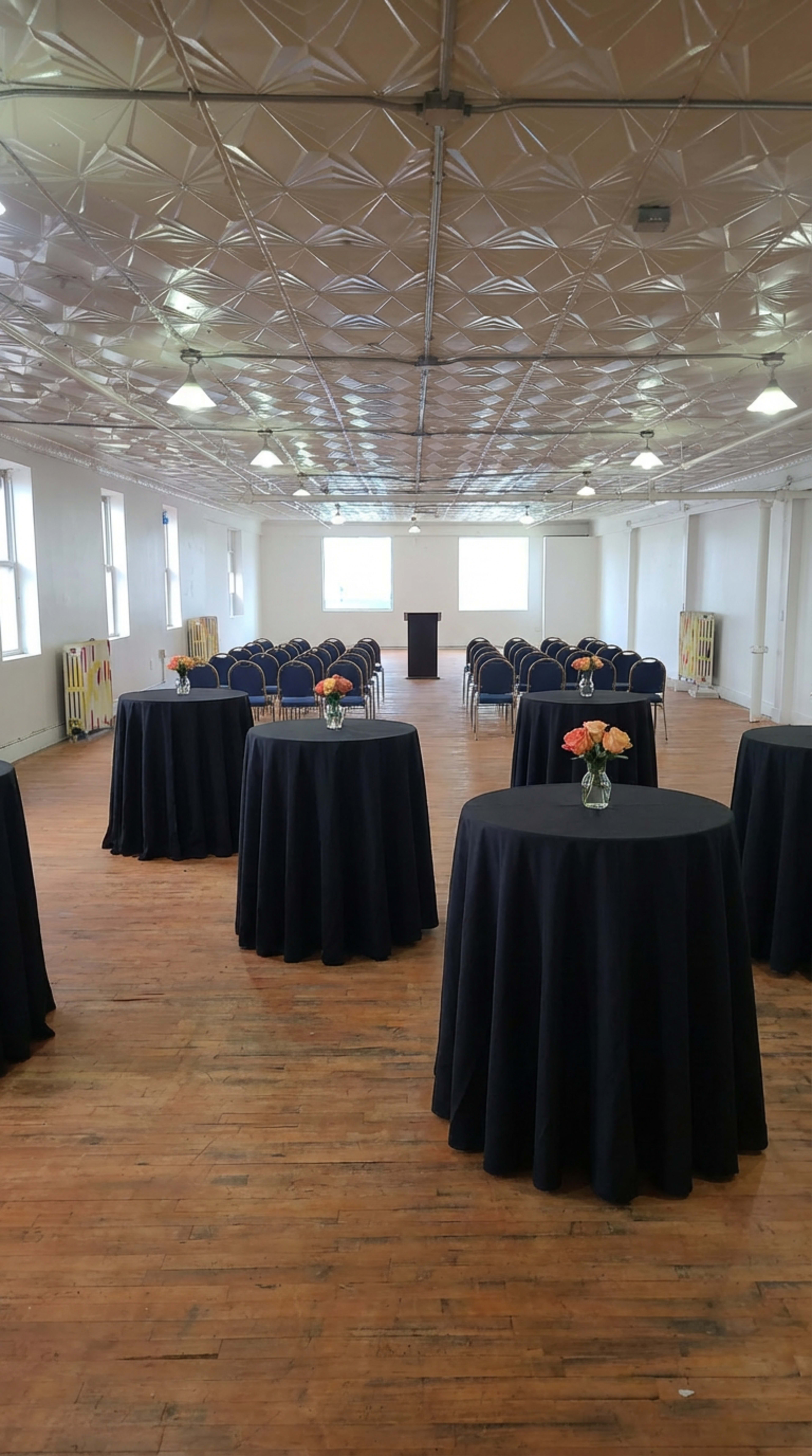 The image shows a large, empty room with several round tables covered in black tablecloths and rows of chairs arranged for an event.