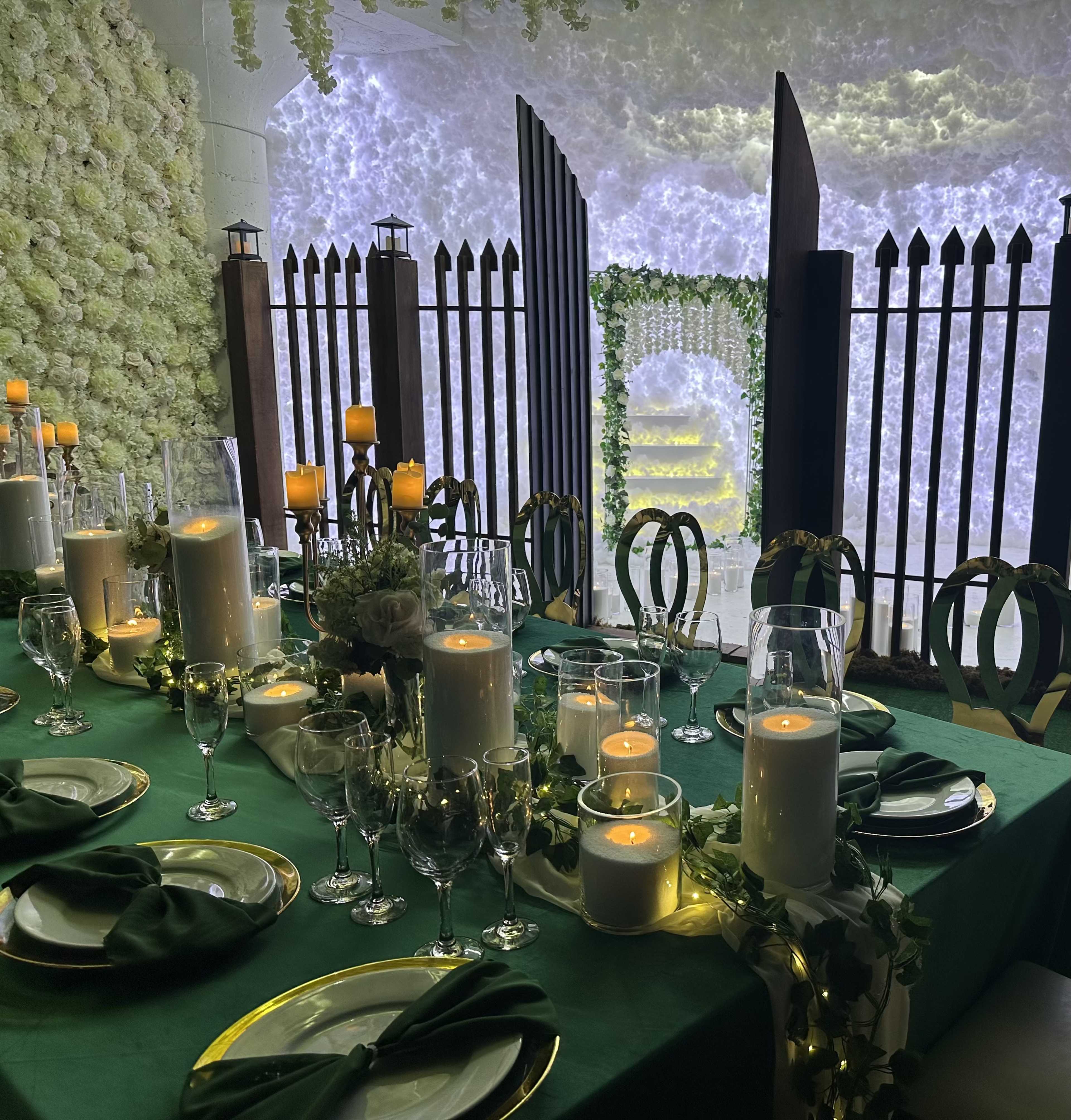 A long dining table is set with green linens, white candles, and decorative glassware, against a backdrop of a floral wall and an illuminated archway.