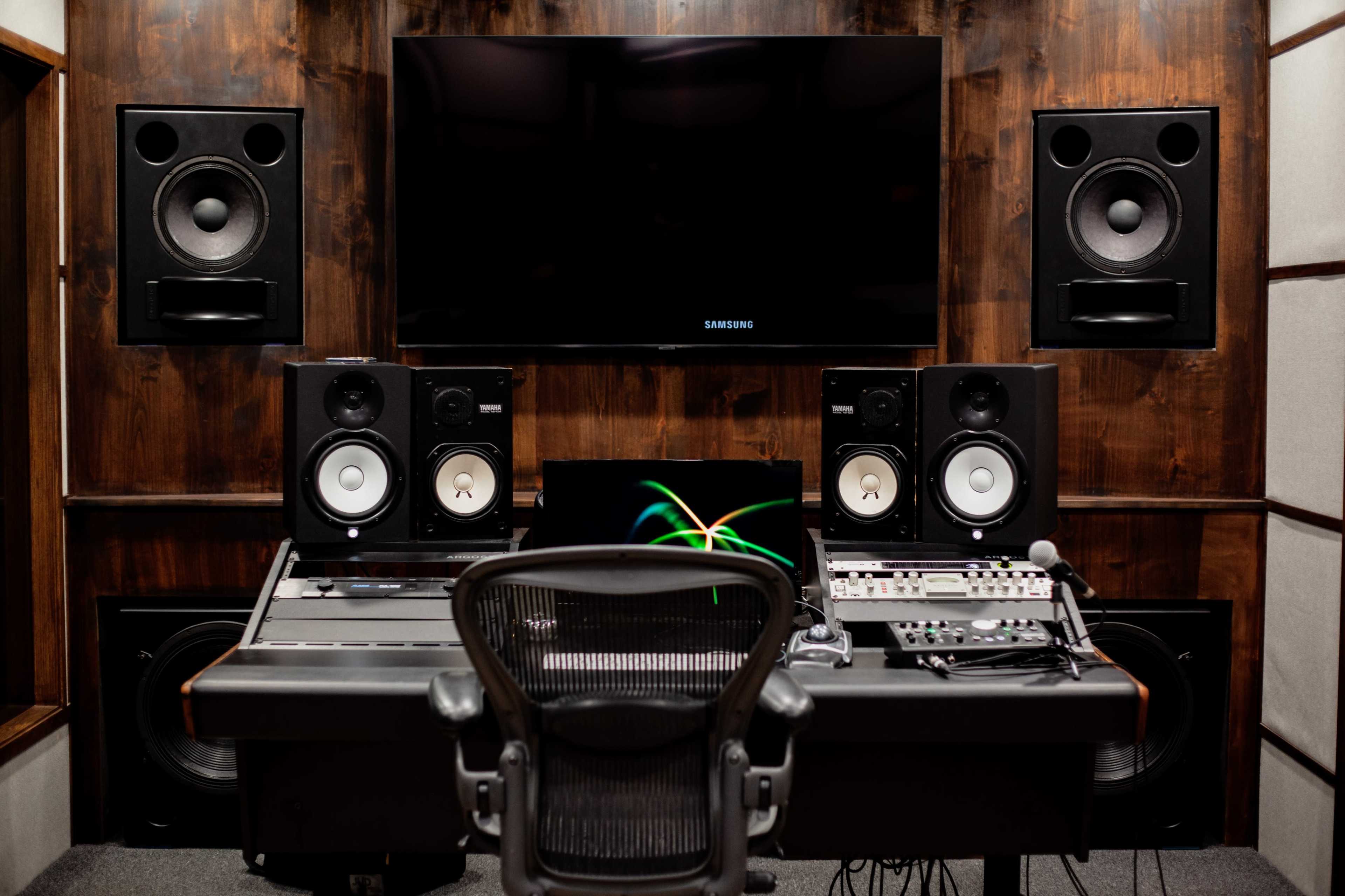 A music studio with a mixing console, two large speaker monitors, and a wall-mounted television, all set against dark wooden paneling.