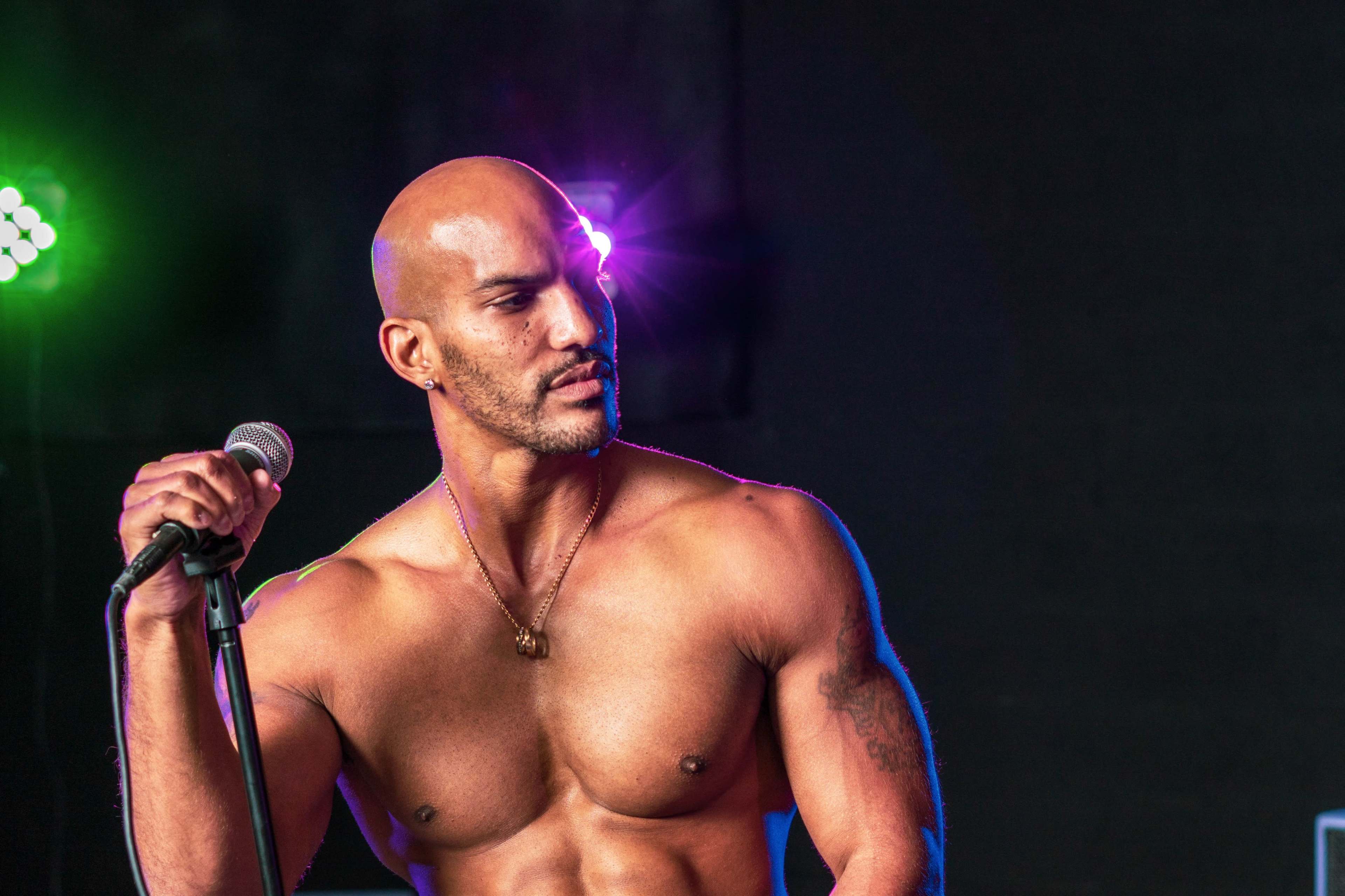 A shirtless man holds a microphone on stage with colorful lights in the background.