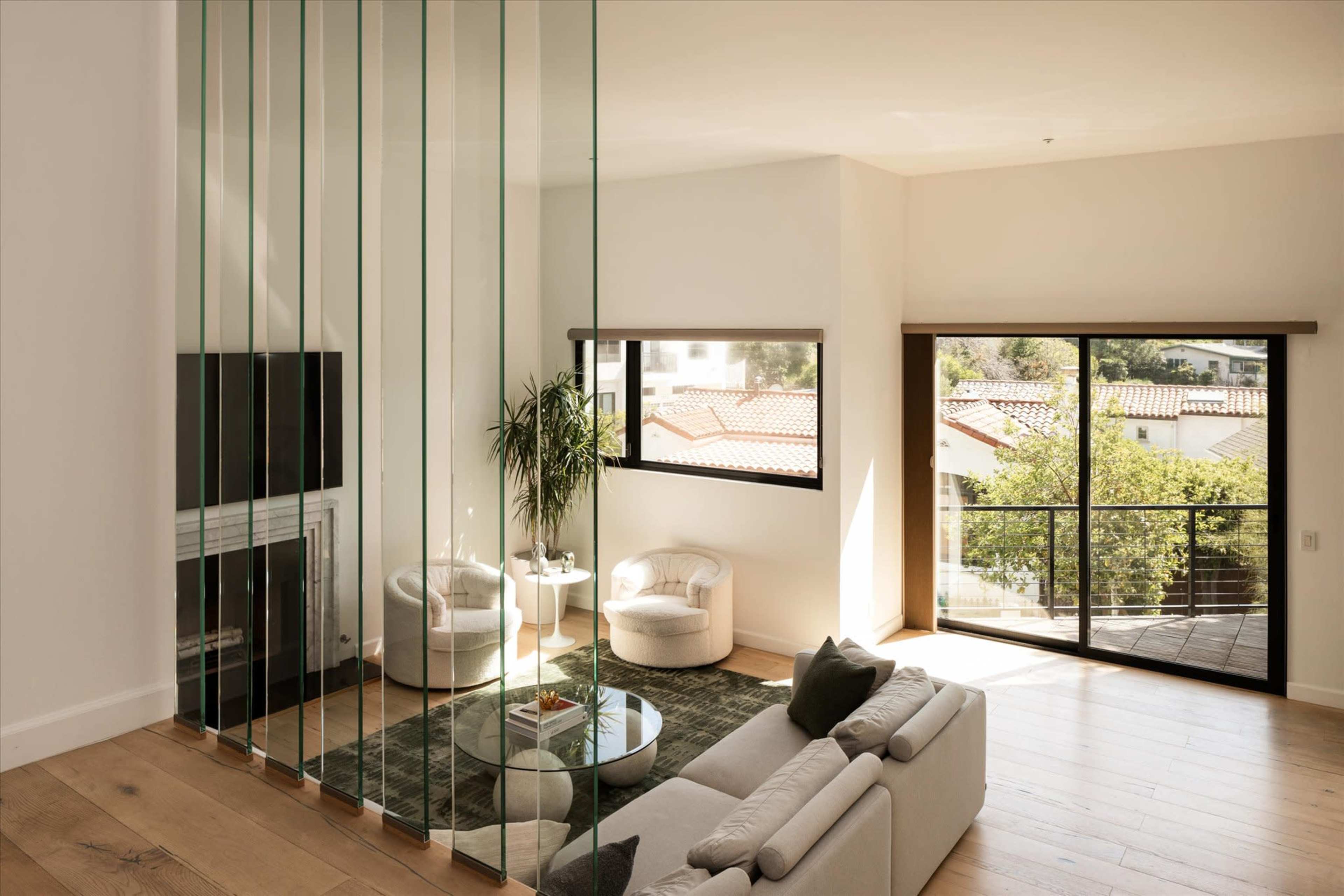 The image shows a modern living room featuring a glass divider, two armchairs, a circular table, and large windows that let in natural light.