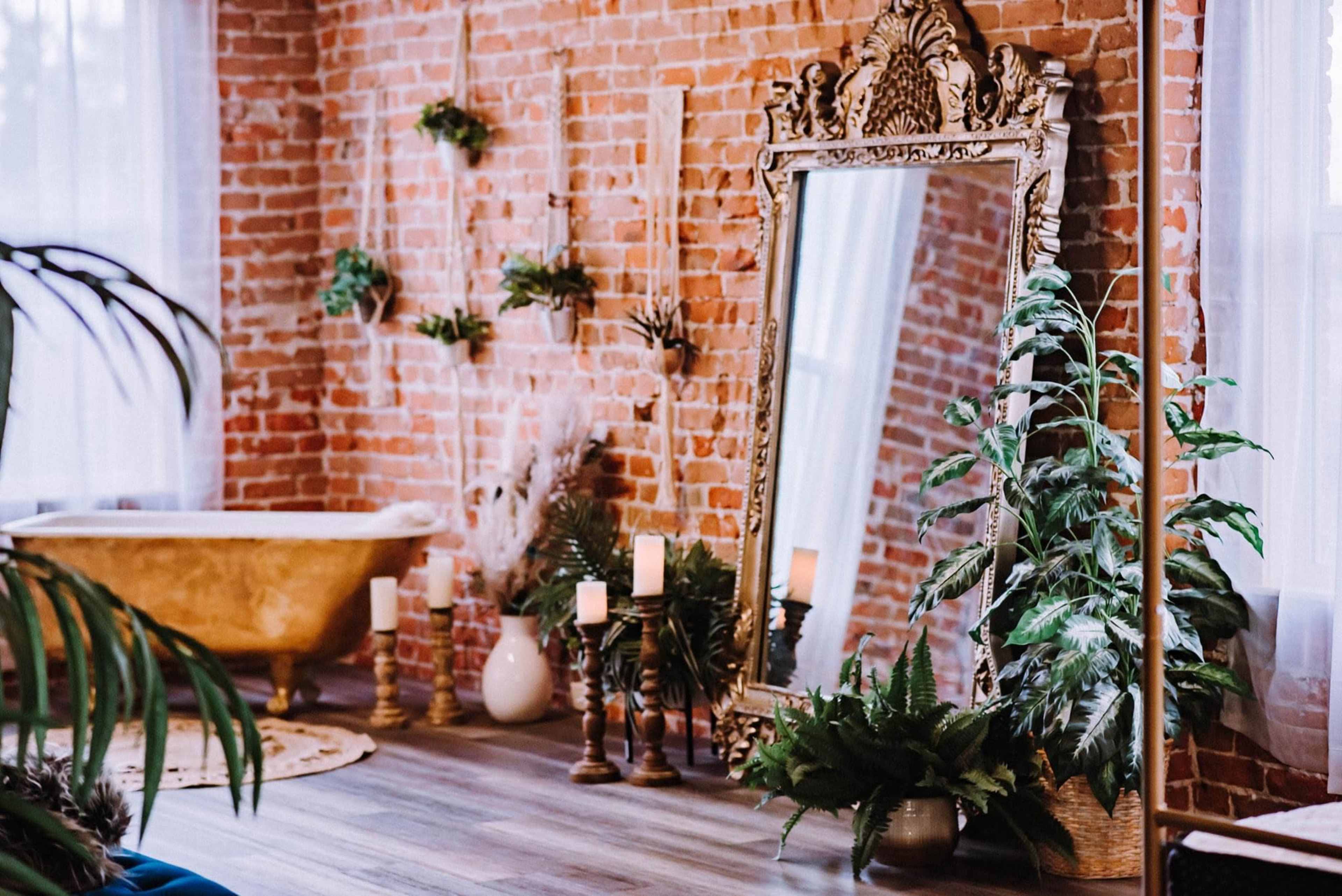 A rustic bathroom with exposed brick walls, a large decorative mirror, a golden bathtub, and various plants arranged around candles.