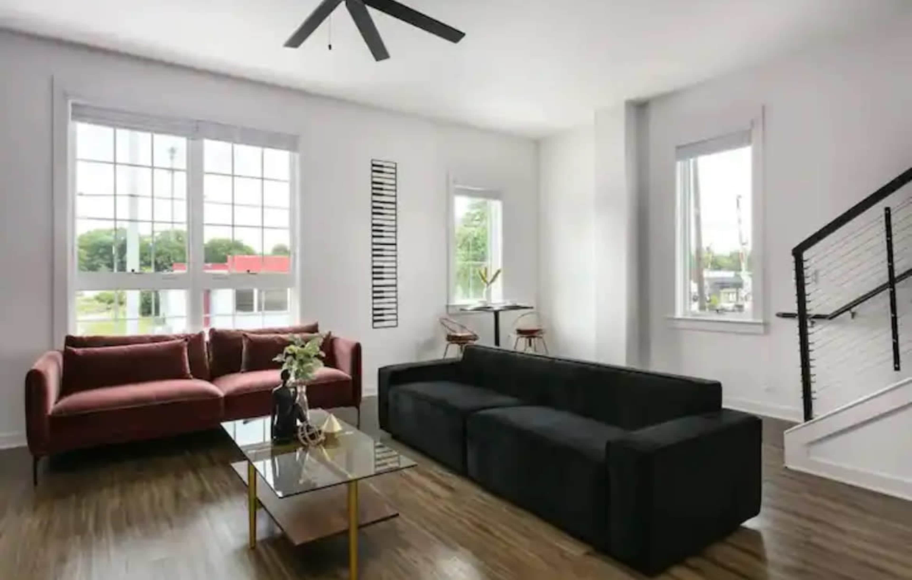 A bright living room featuring a black sofa, a maroon couch, a glass coffee table, and large windows with a staircase in the background.