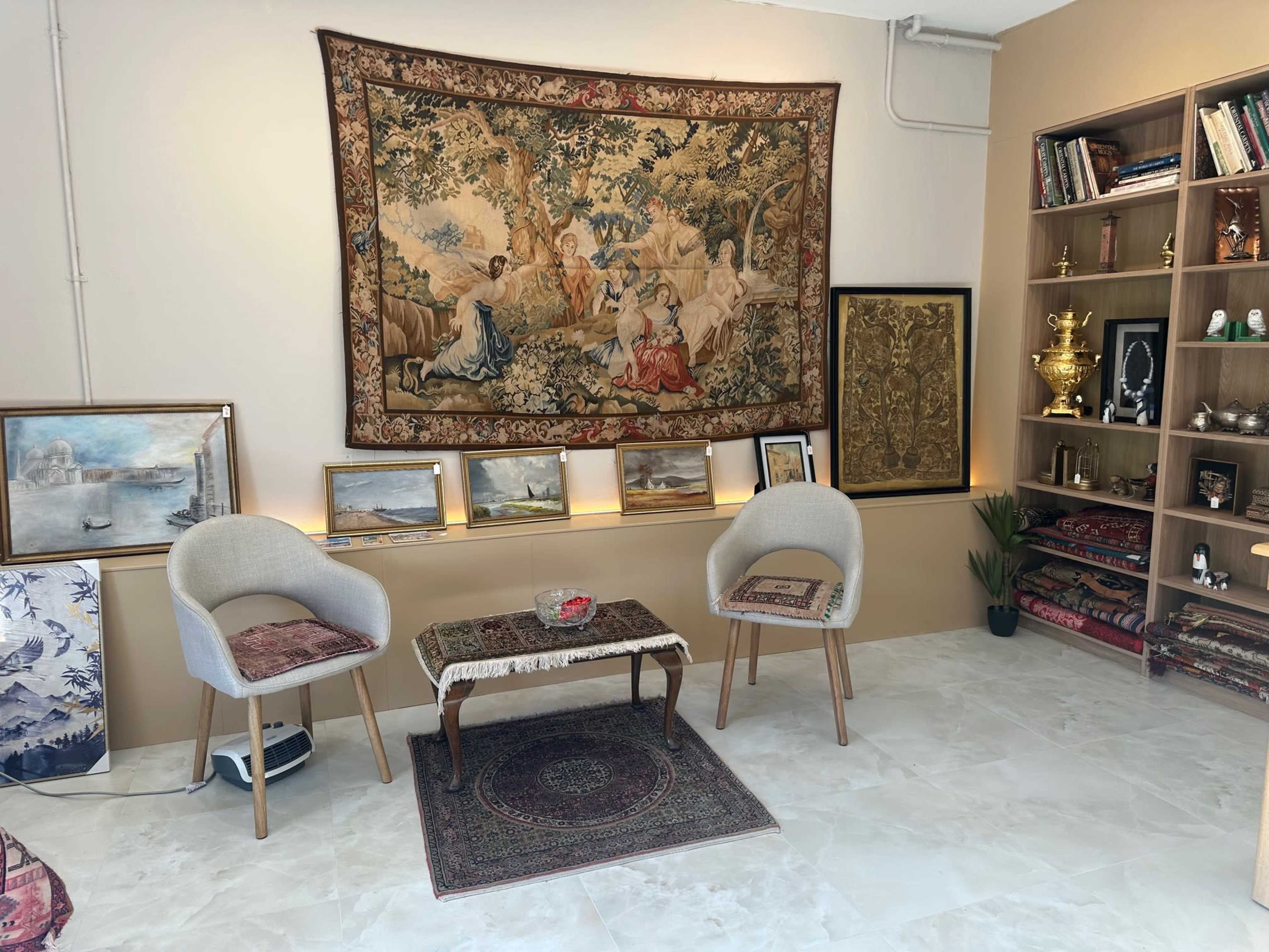 The image shows a cozy room featuring two cream-colored chairs, a small coffee table, decorative wall art, and shelves filled with books and decorative items.