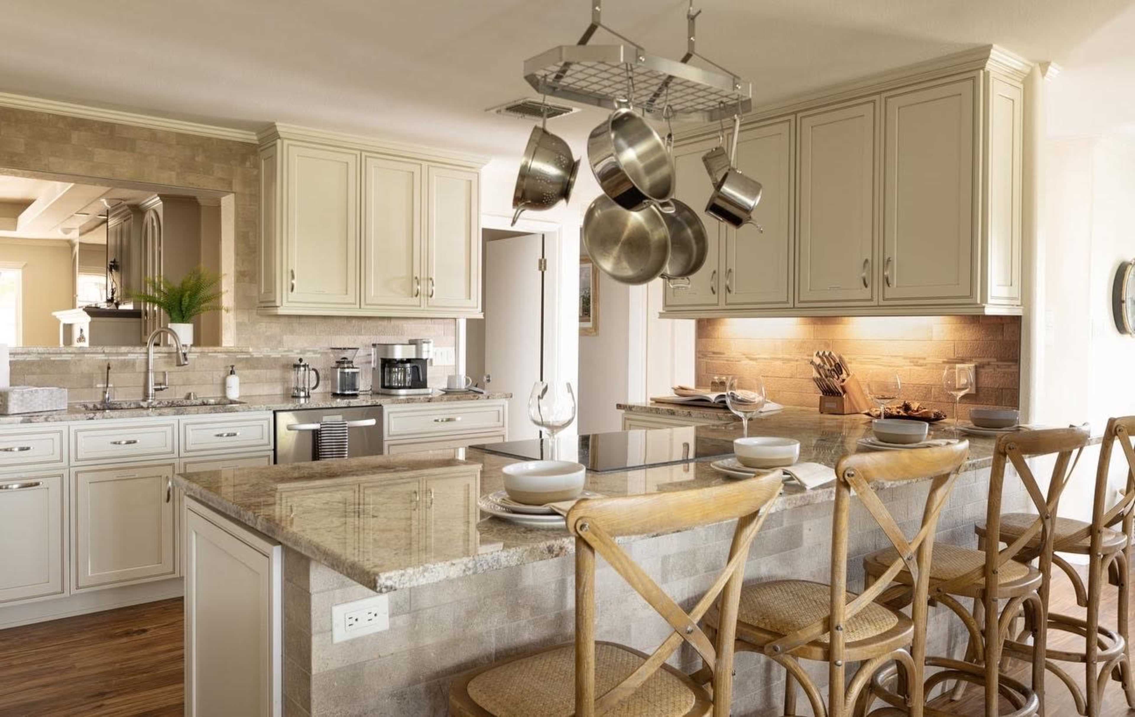 A modern kitchen features cream-colored cabinets, a large island with bar stools, and hanging cookware over a marble countertop.