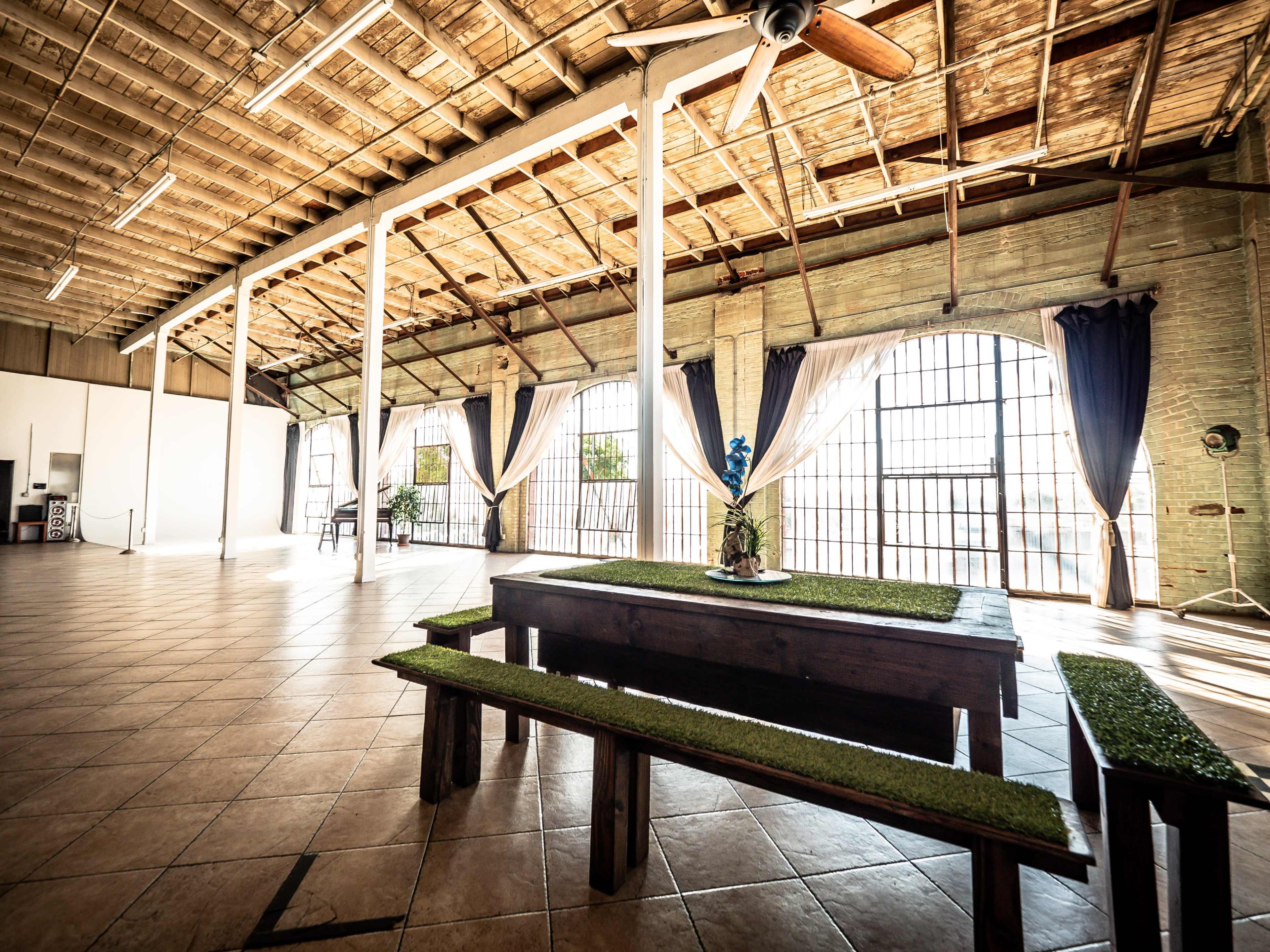 A spacious interior with high ceilings, large windows, and a central wooden table surrounded by benches, all set on tiled flooring.