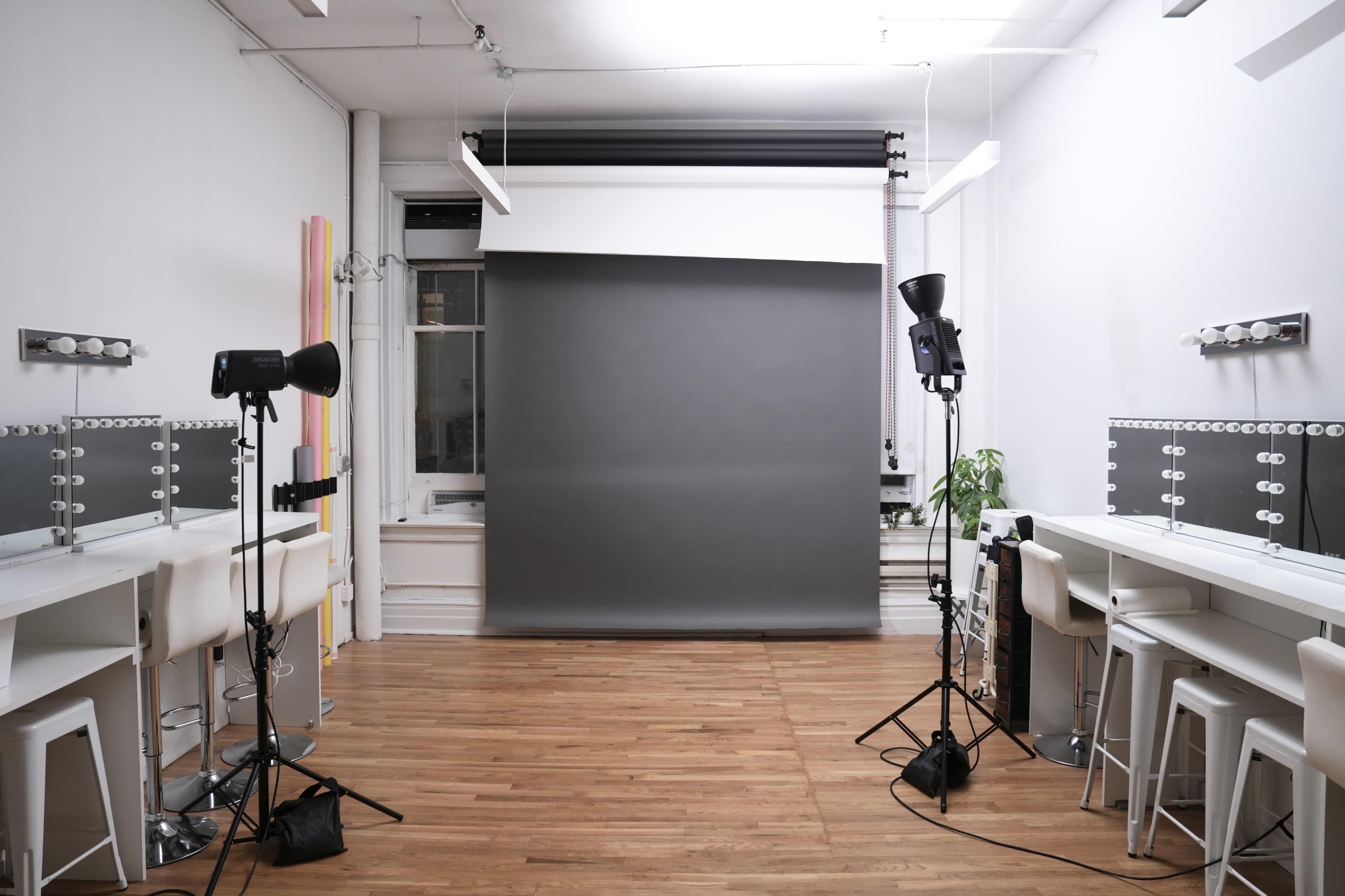 The image shows a photography studio equipped with two lighting setups, several makeup stations, and a gray backdrop.
