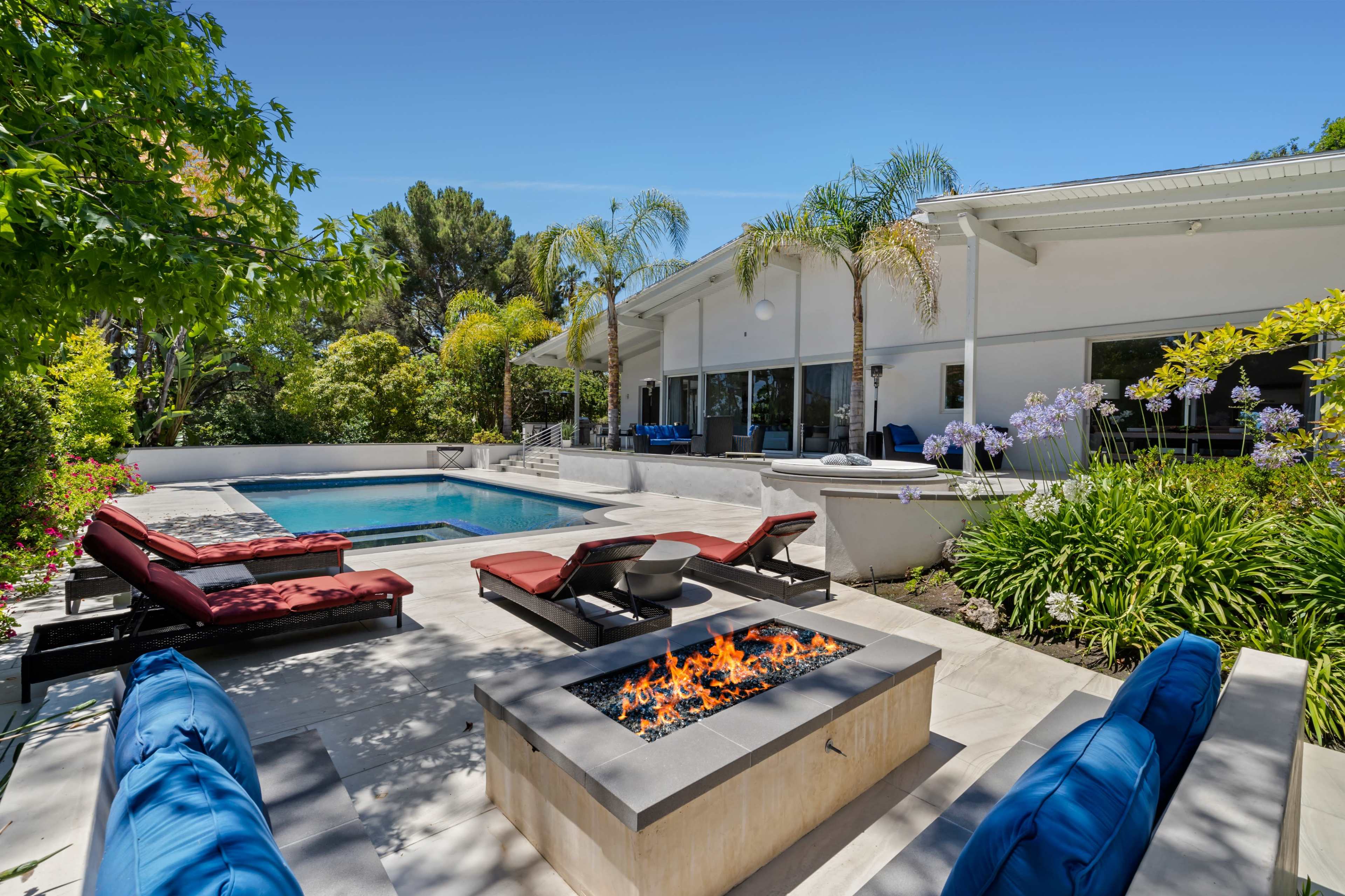 The image shows a modern backyard with a swimming pool, lounge chairs, and a fire pit surrounded by lush greenery.