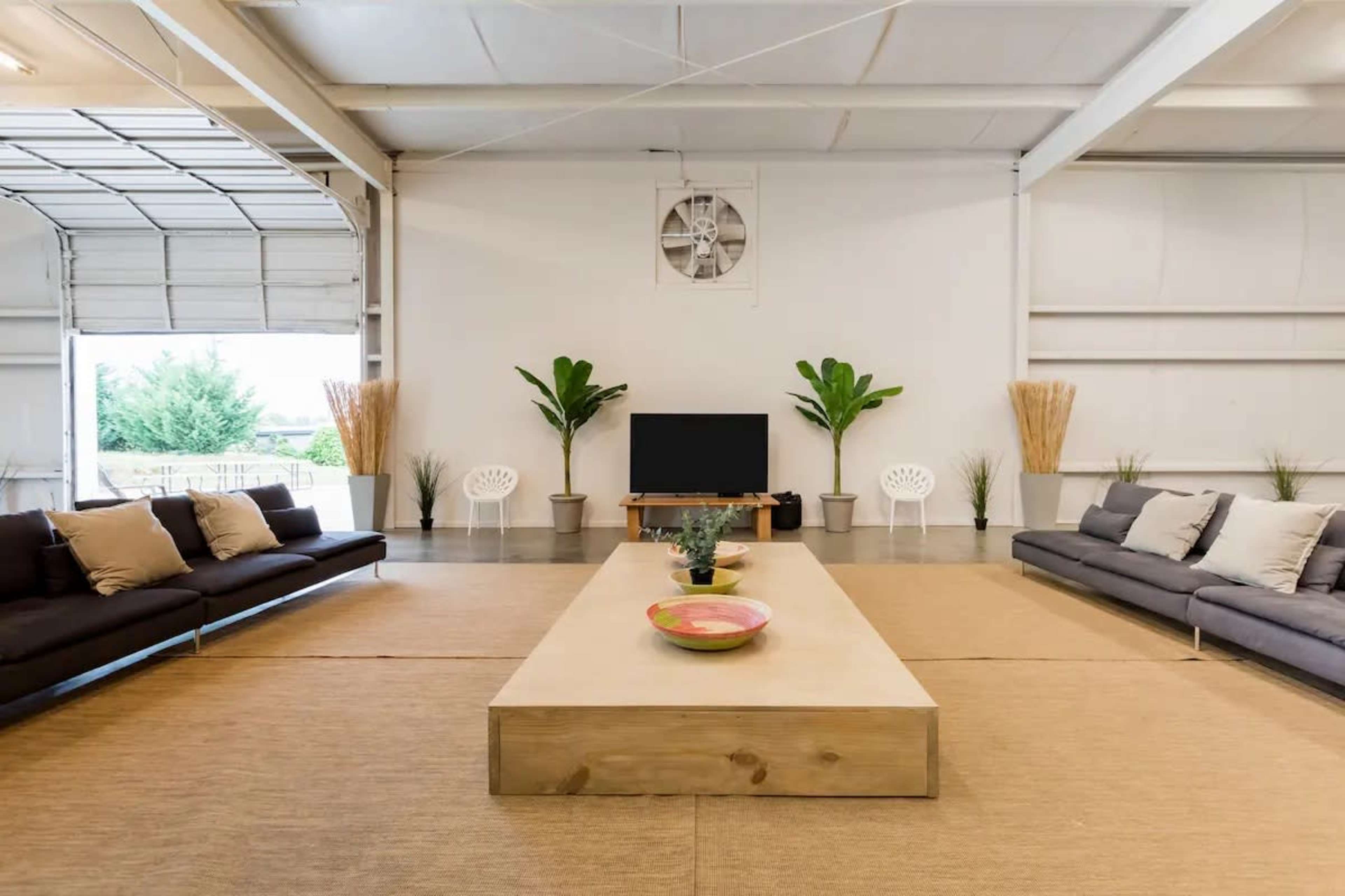The image shows a spacious, modern living area with two dark sofas facing a central wooden coffee table, a television on a stand, and large plants in the corners.