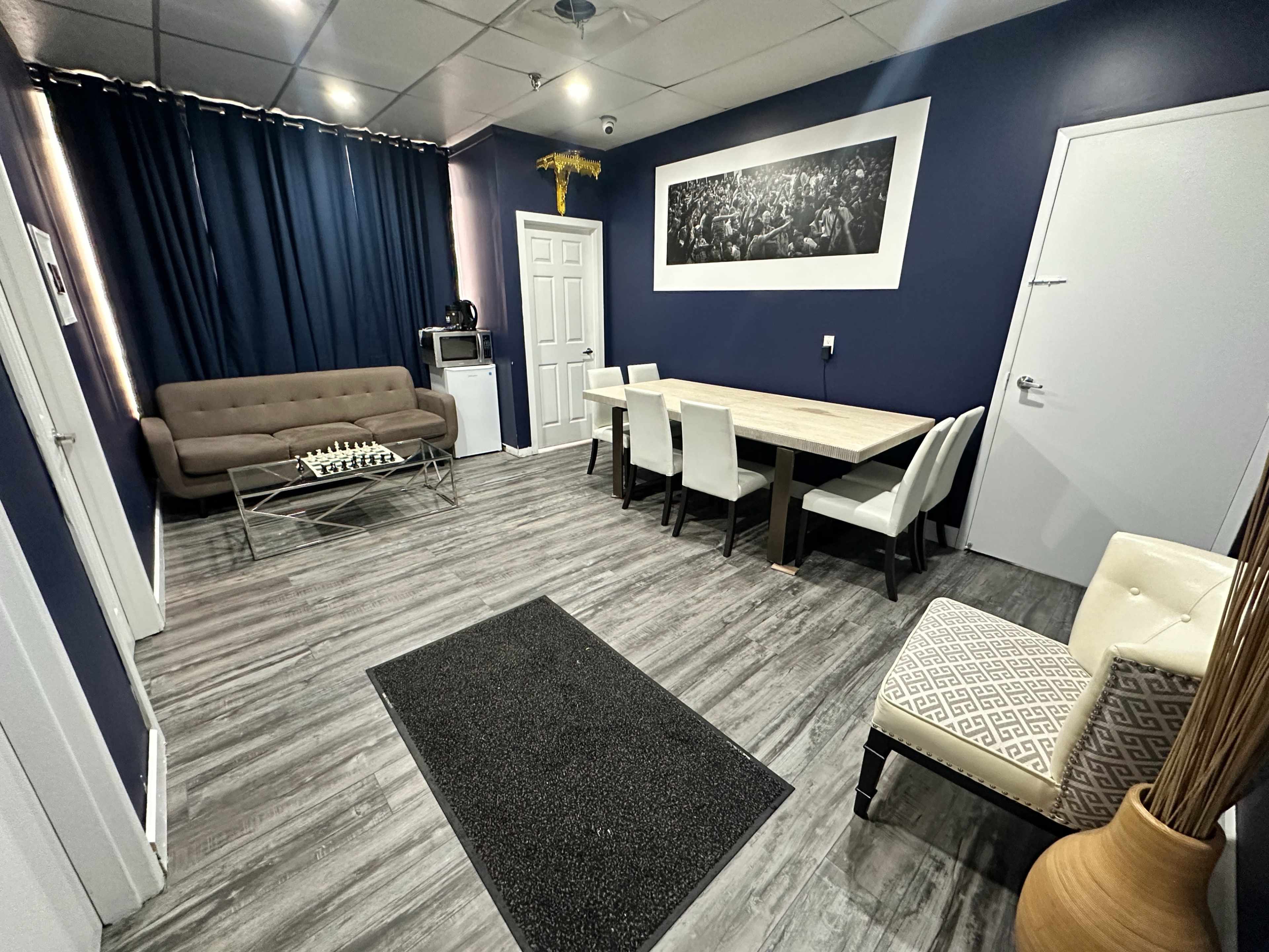 The image shows a modern, minimalist room featuring a sectional sofa, a wooden dining table with chairs, and decorative elements, with dark blue walls and a contrasting floor.