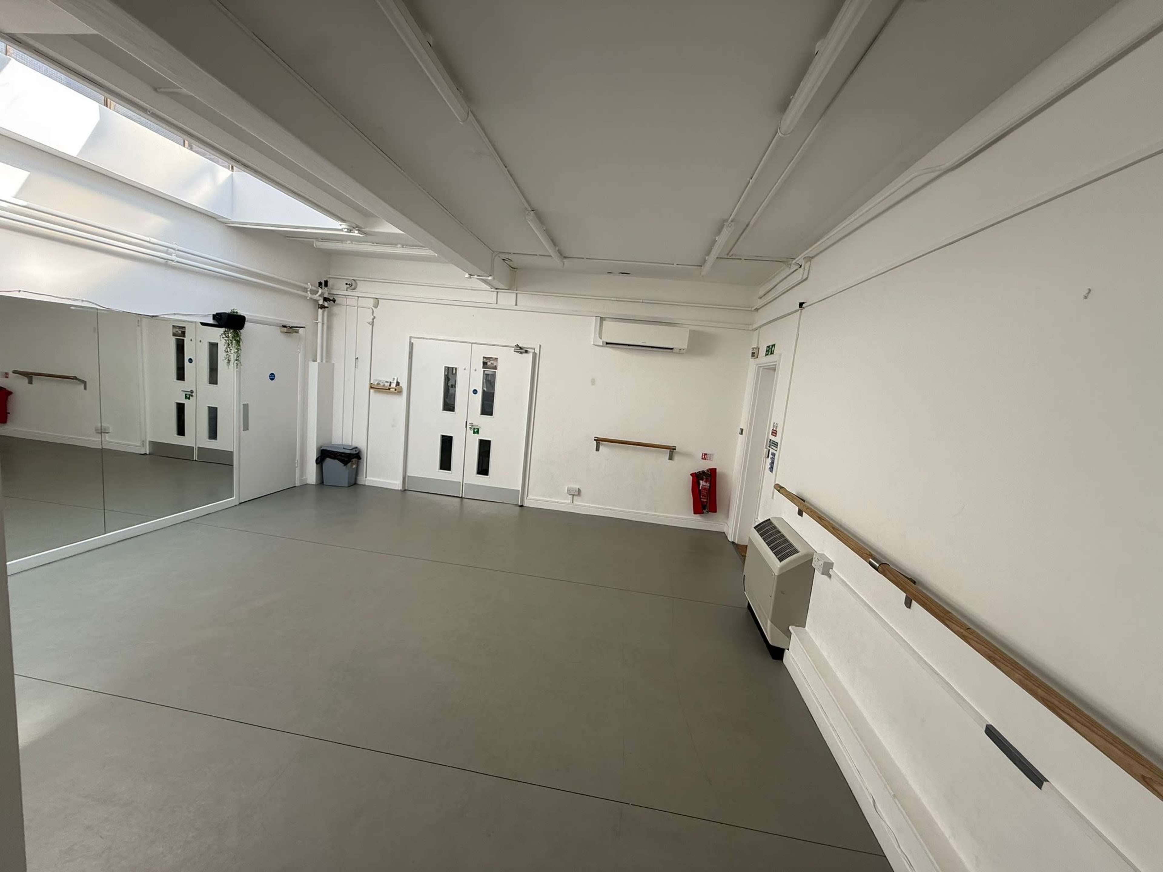 The image shows a spacious, empty studio with a mirrored wall, gray flooring, and overhead lighting.