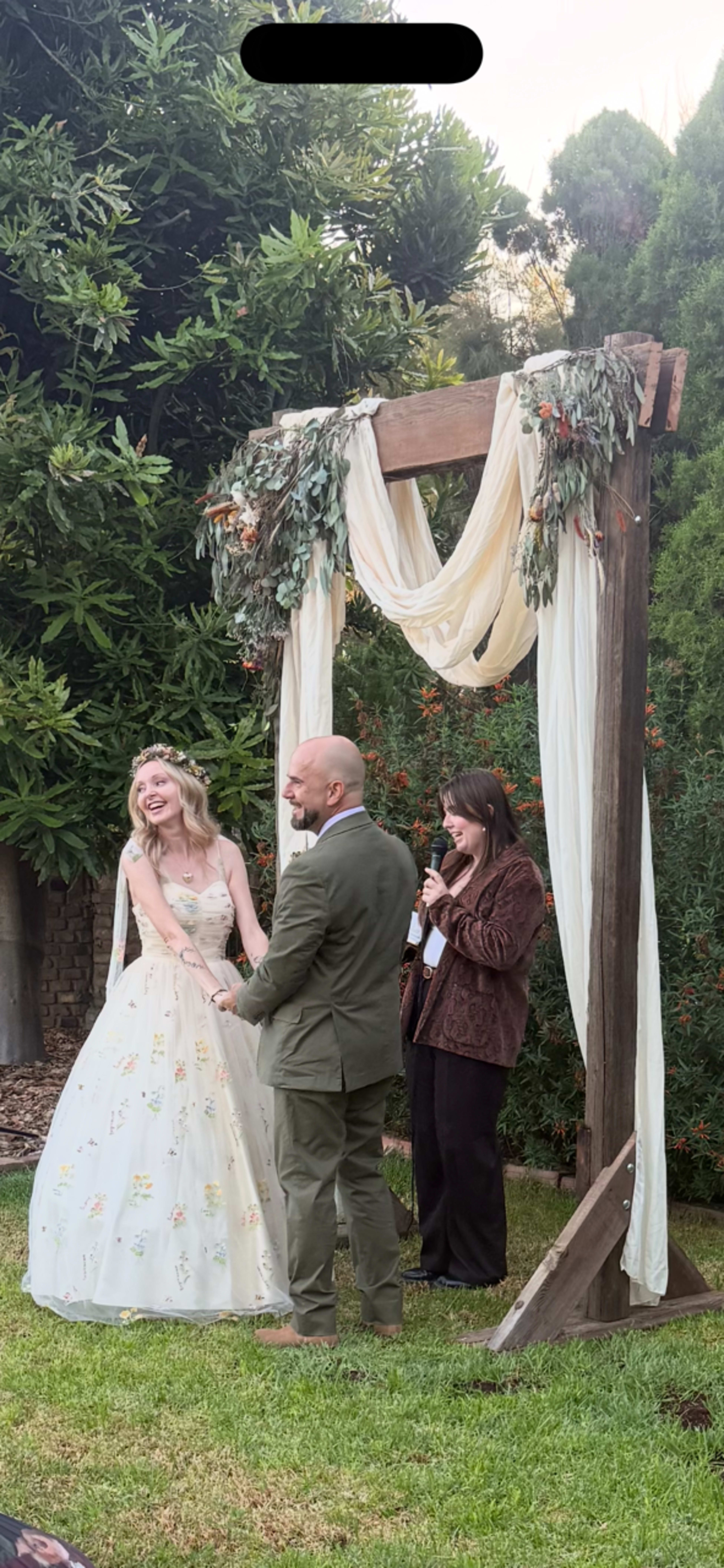 A couple stands in front of a floral-decorated arch while exchanging vows during an outdoor wedding ceremony.
