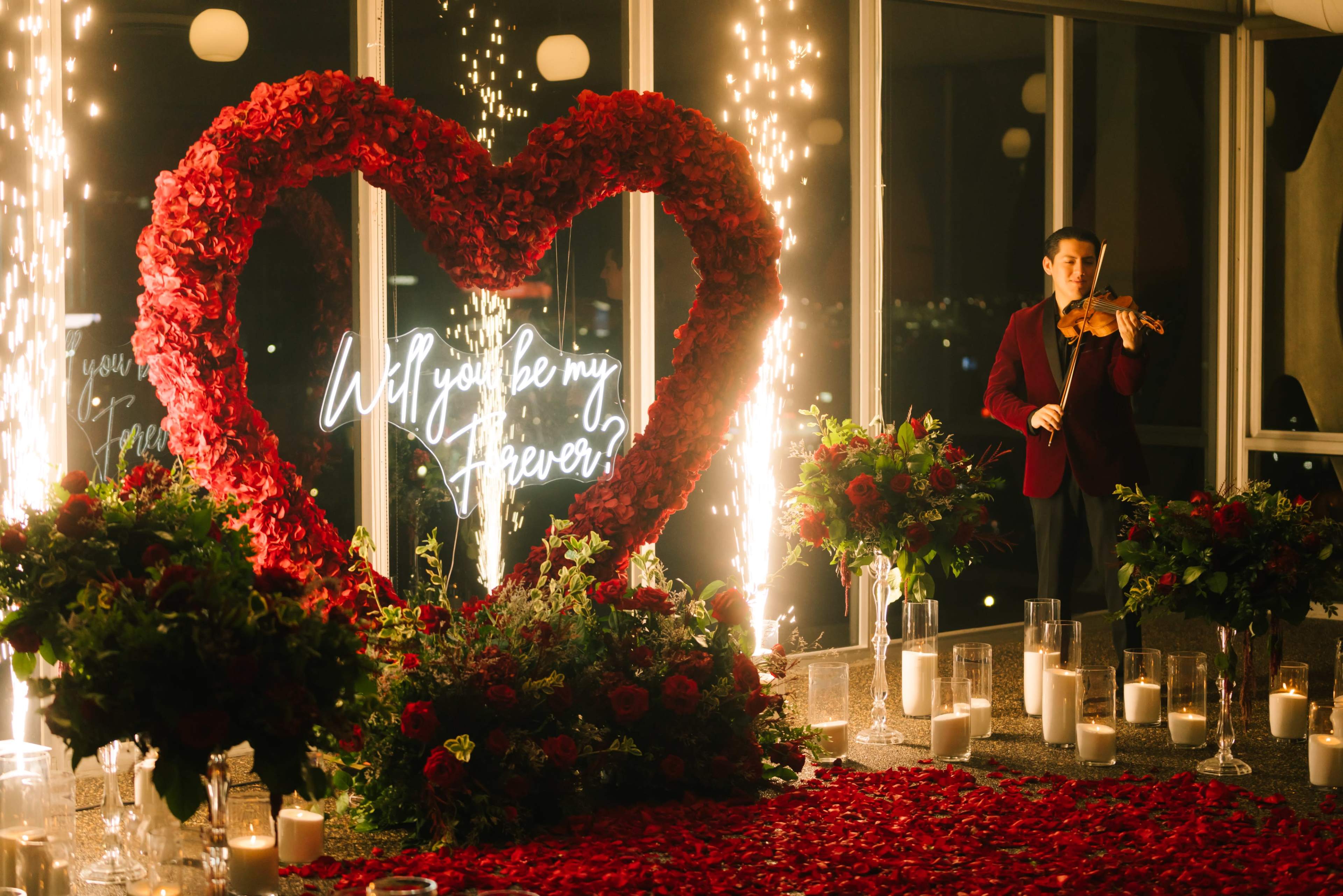 A violinist stands next to a large heart made of red flowers, surrounded by candles and festive decorations, while a neon sign reads "Will you be my Forever?" in a warmly lit setting.