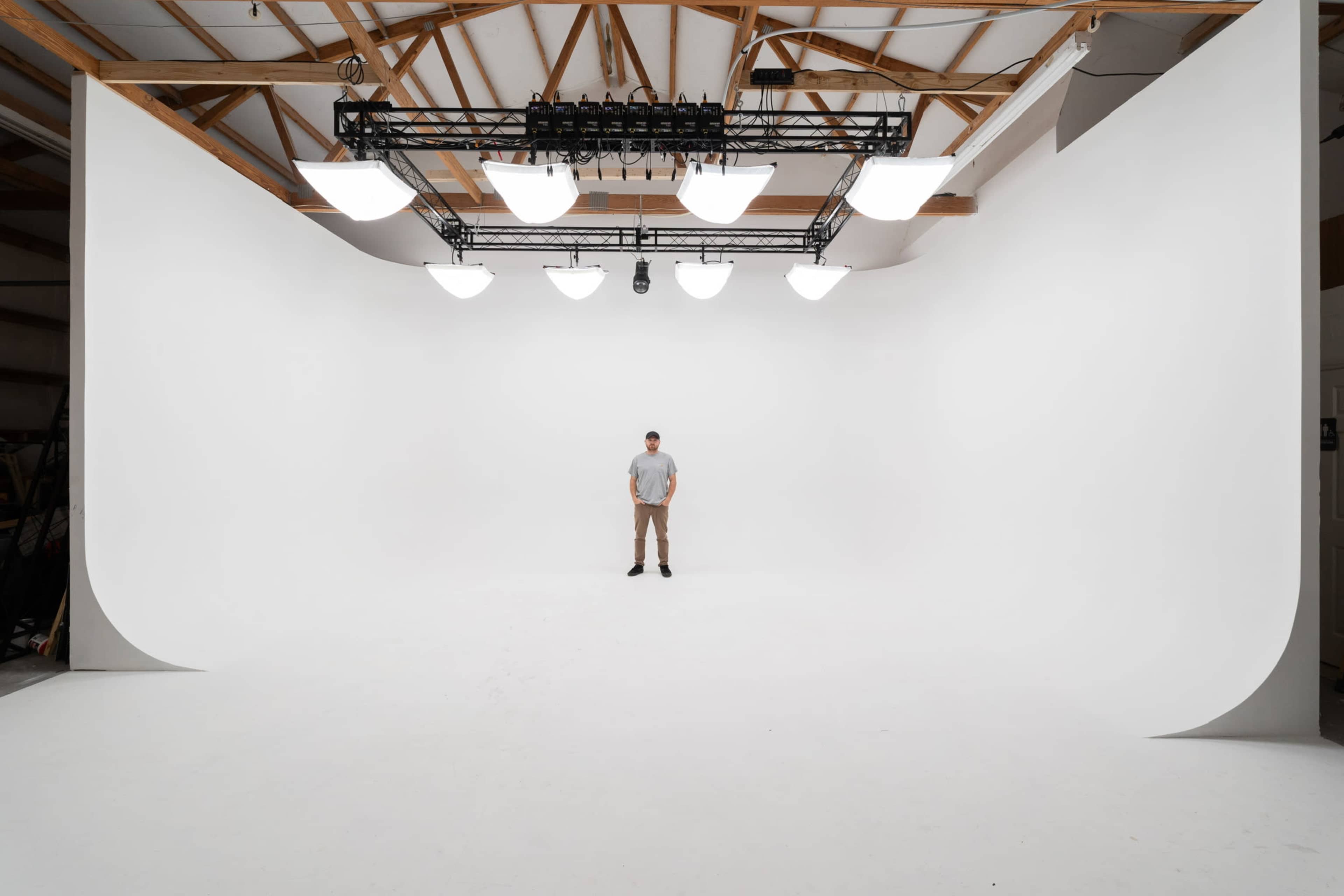 A person stands alone in a spacious studio with a seamless white backdrop and overhead lighting.