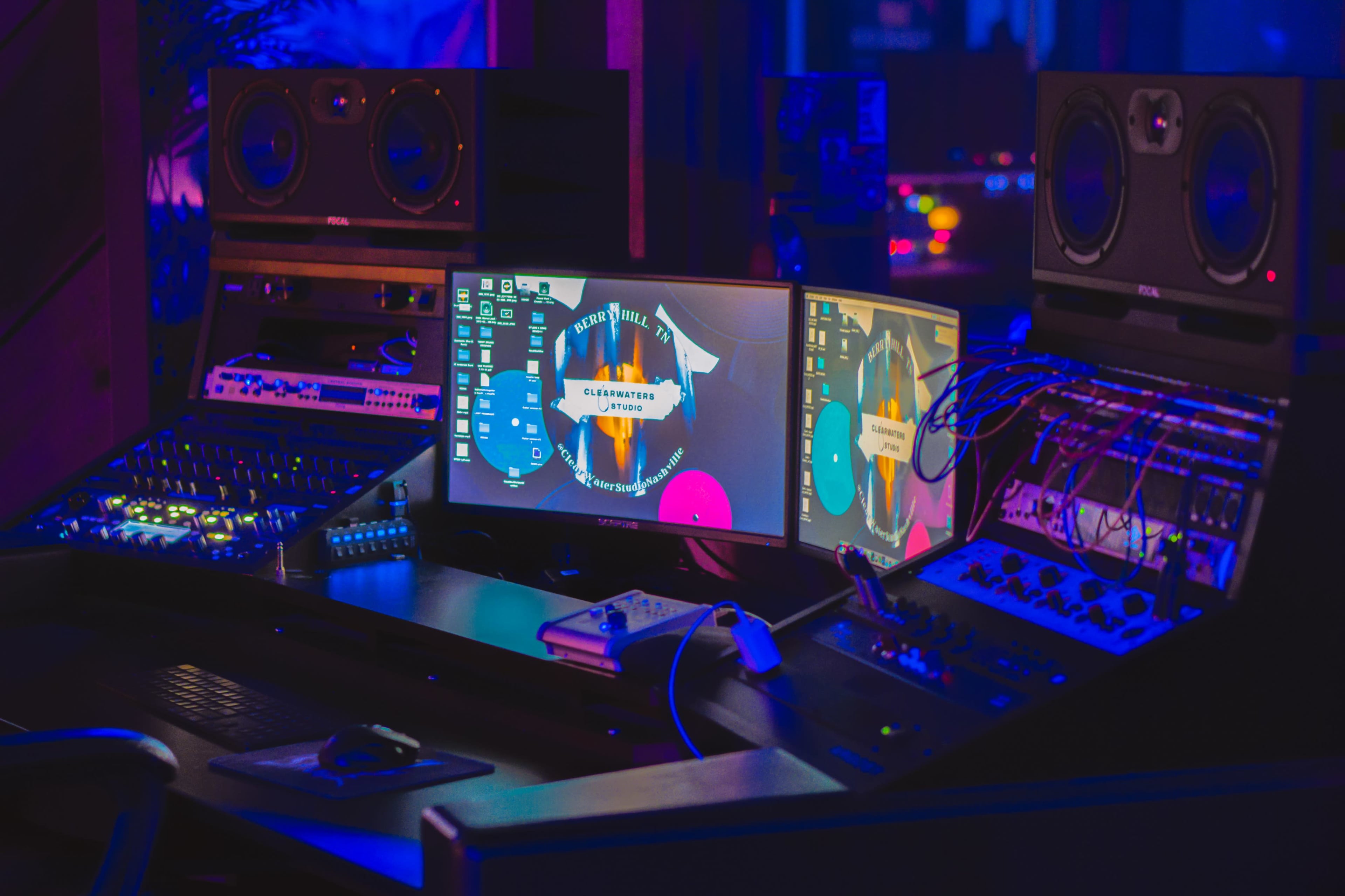 A computer workstation in a music studio features multiple monitors, audio equipment, and a digital interface amidst a blue-lit environment.