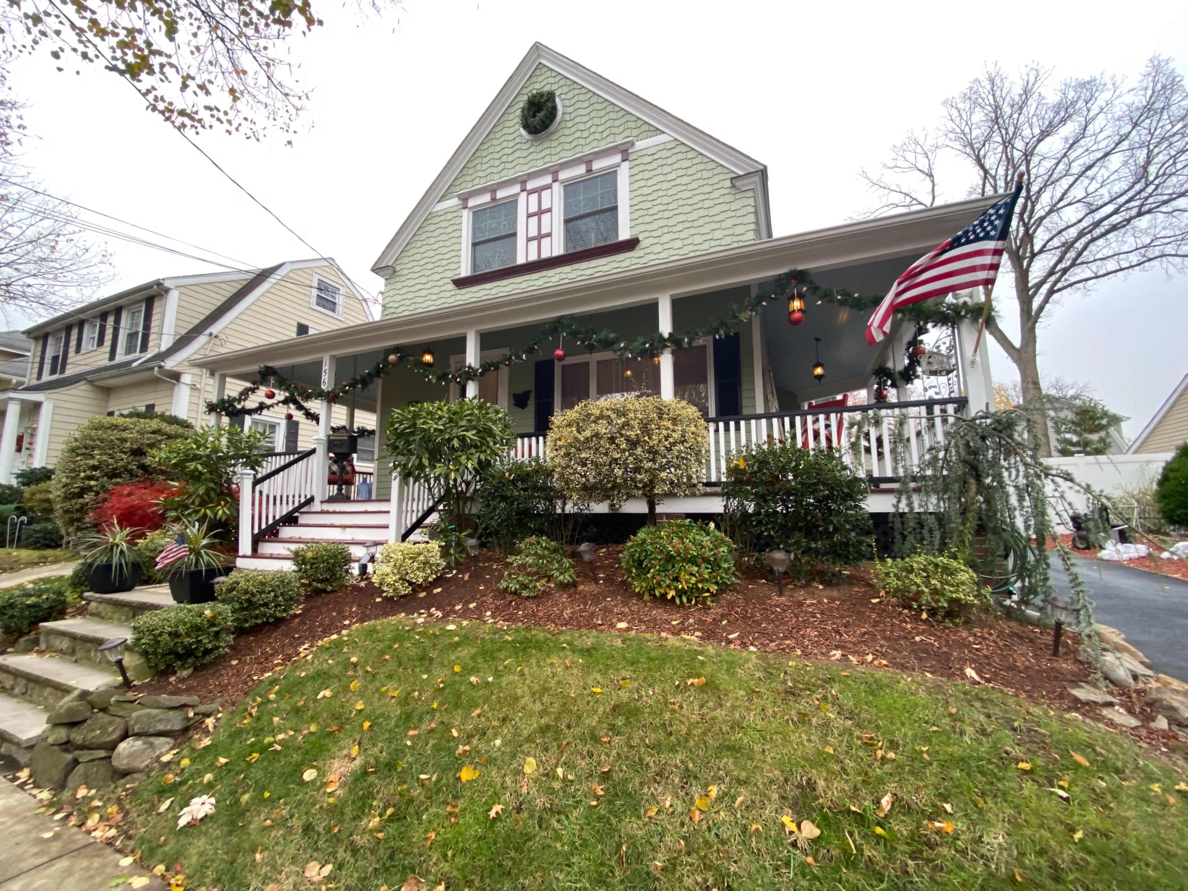 A green, two-story house with a front porch is decorated for the holidays, featuring garlands, ornaments, and an American flag.