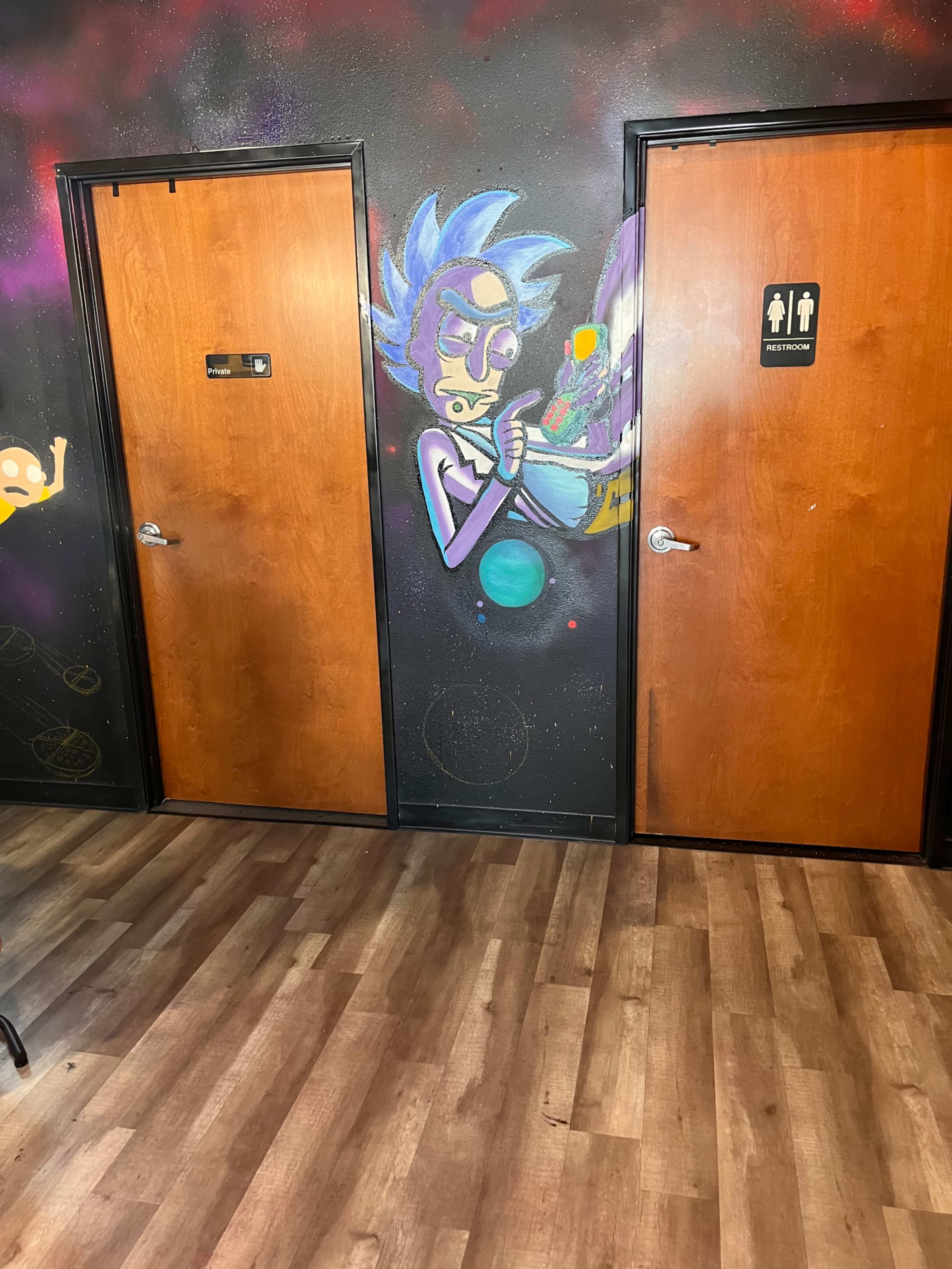 The image depicts a wall with two wooden doors, one labeled with a symbol for restrooms, alongside a colorful mural of a character holding a device.