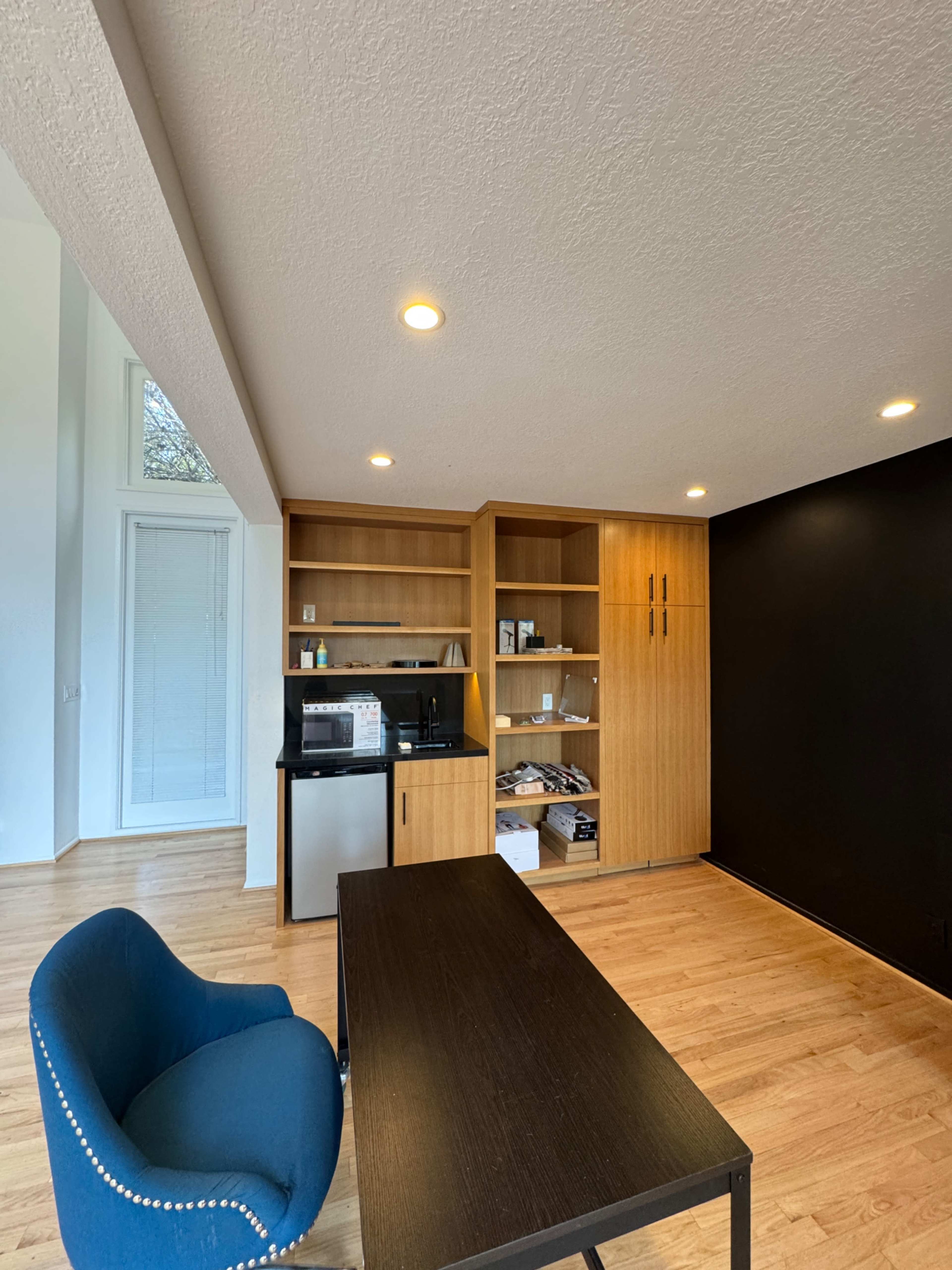 A modern room features a black accent wall, a wooden shelving unit with a microwave and mini fridge, and a blue chair beside a black table.