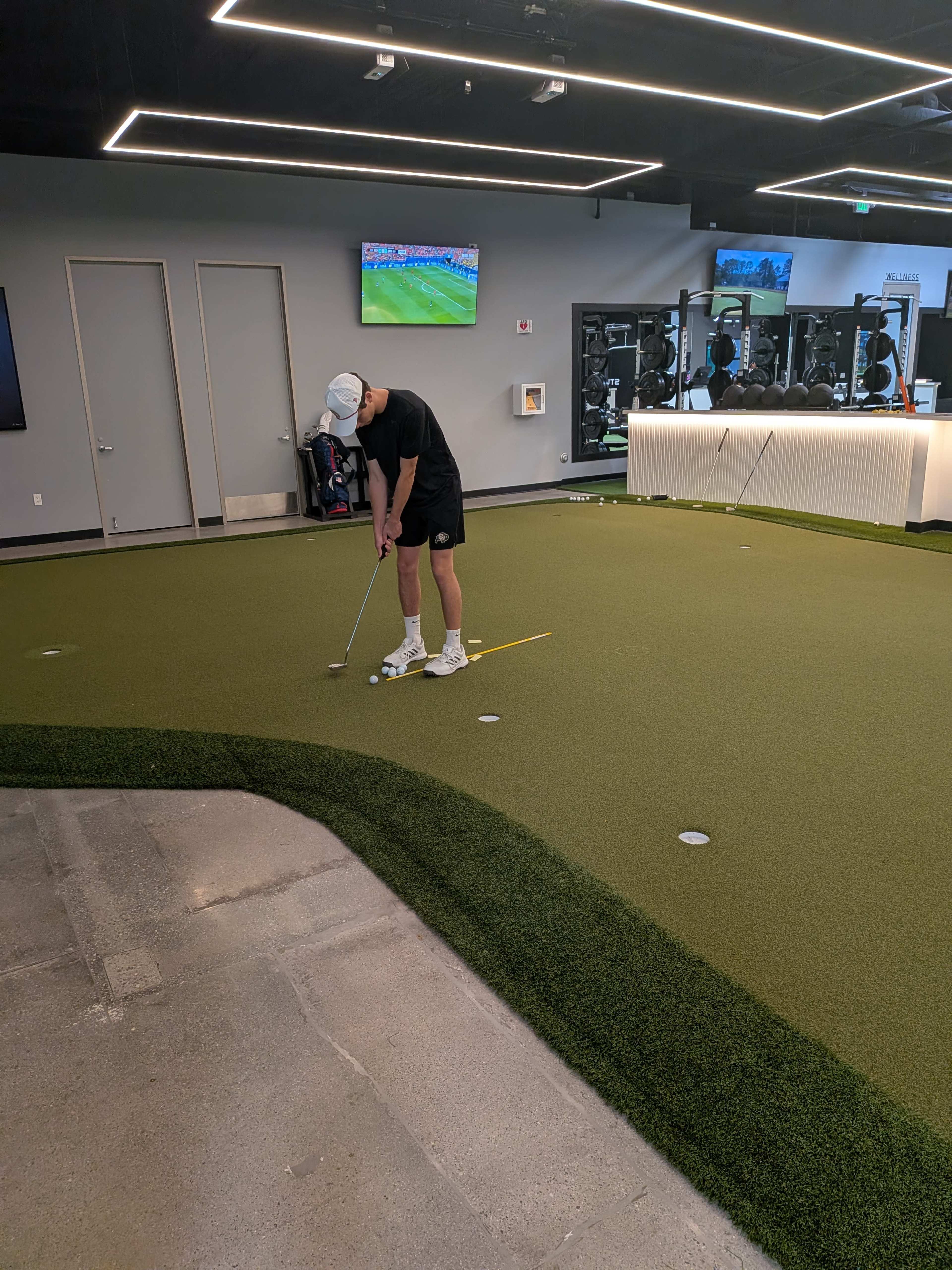 A person is practicing putting on a synthetic green surface in a gym-like setting with exercise equipment and televisions in the background.