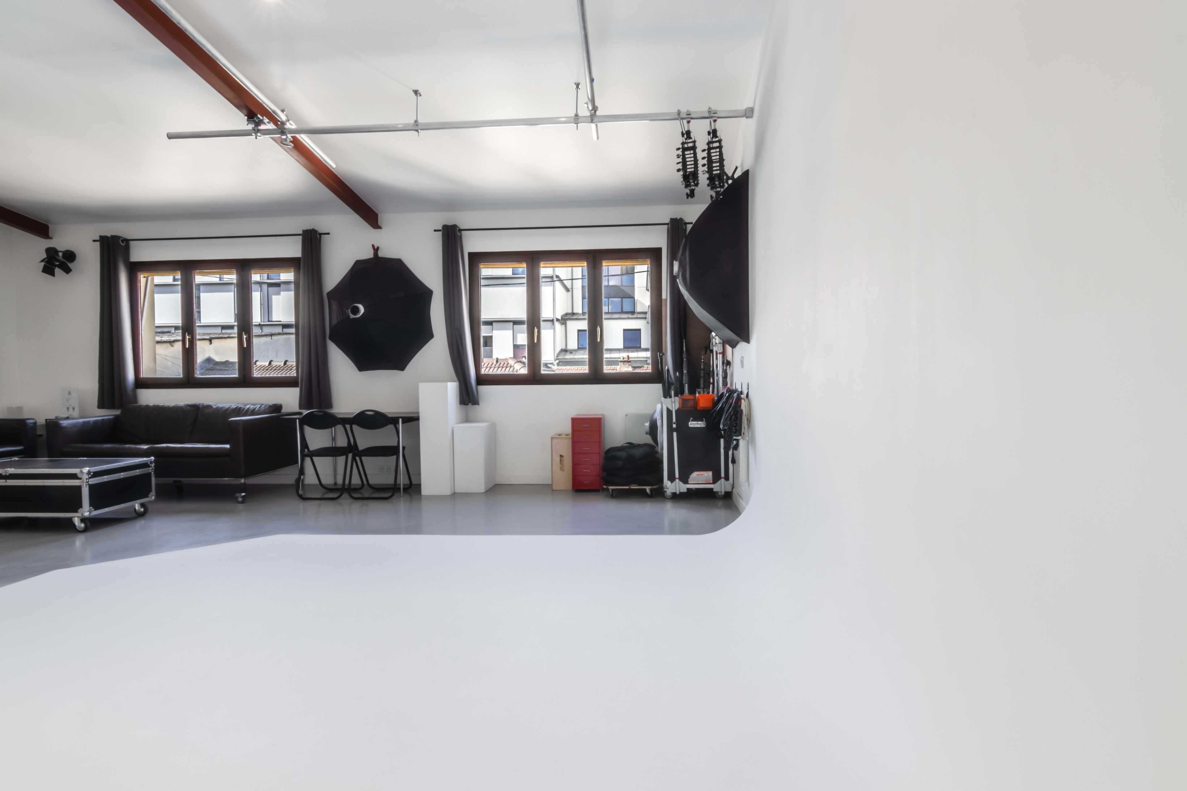 A photography studio featuring a curved white backdrop, a black sofa, a set of chairs, and various lighting equipment.