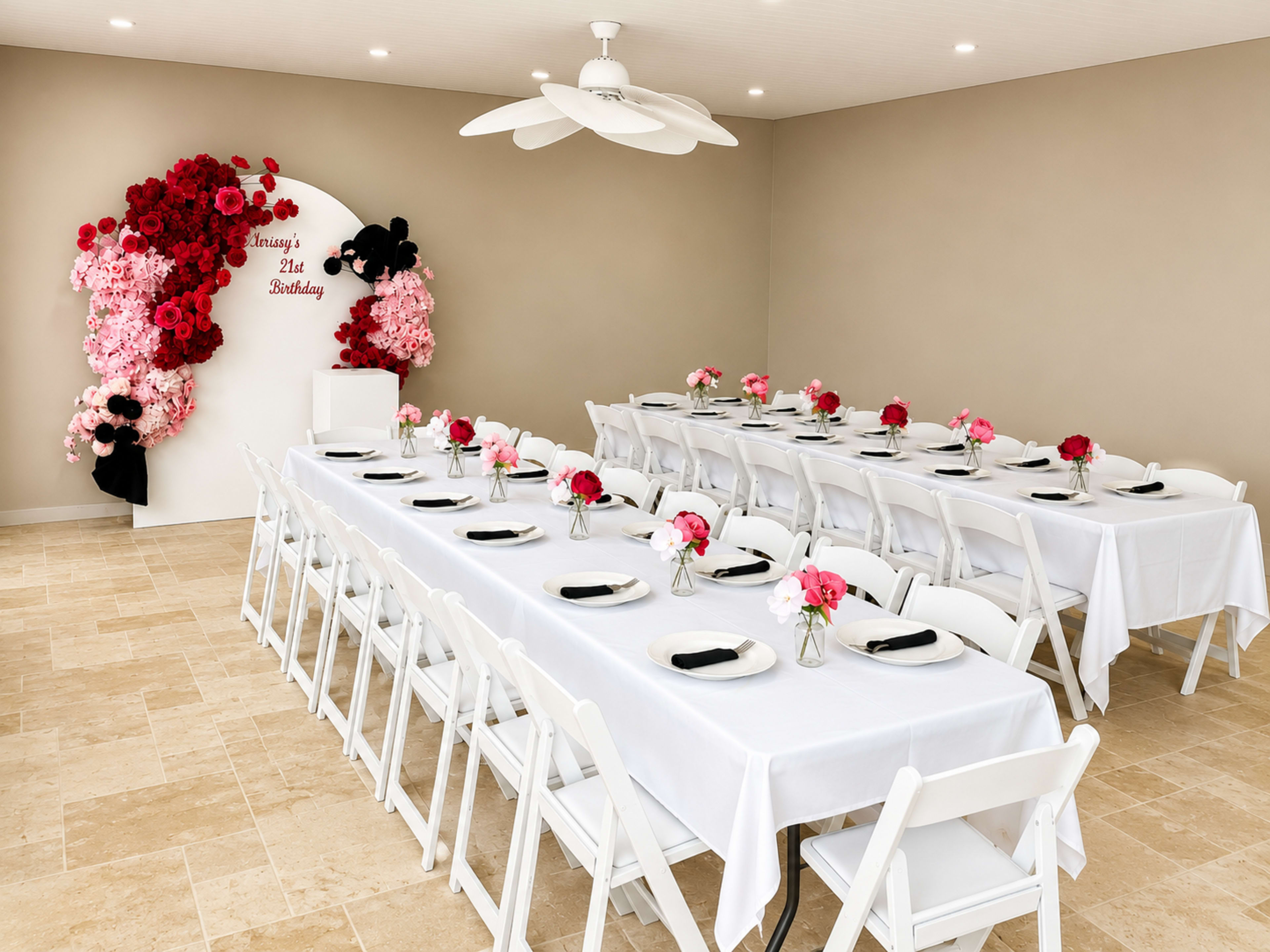 A large, decorated room features two long tables set with white tablecloths, black plates, and floral centerpieces, along with a backdrop of red and pink flowers for a birthday celebration.