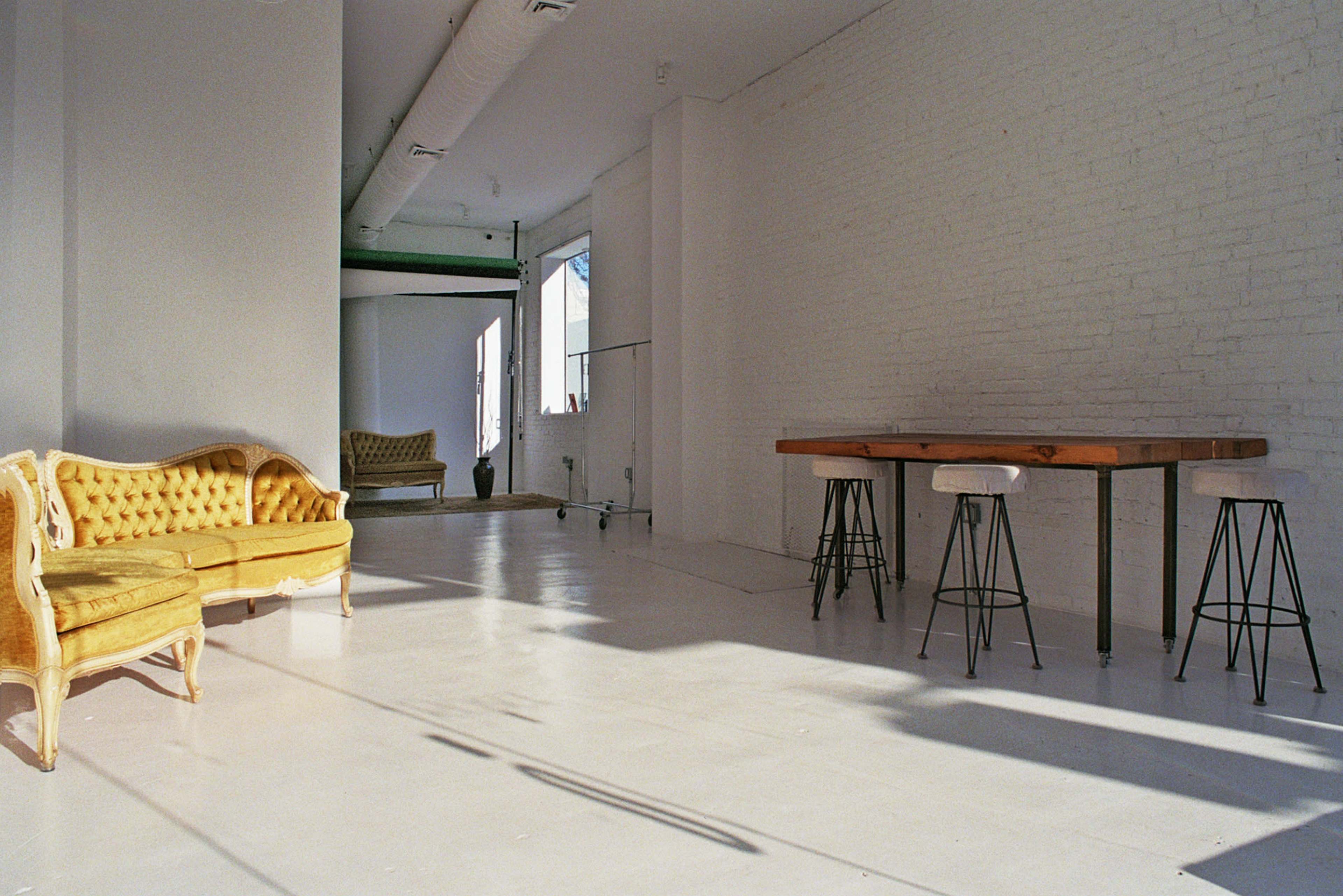 A bright, minimalist indoor space features a vintage yellow sofa, a wooden table with metal stools, and large windows allowing natural light to fill the room.