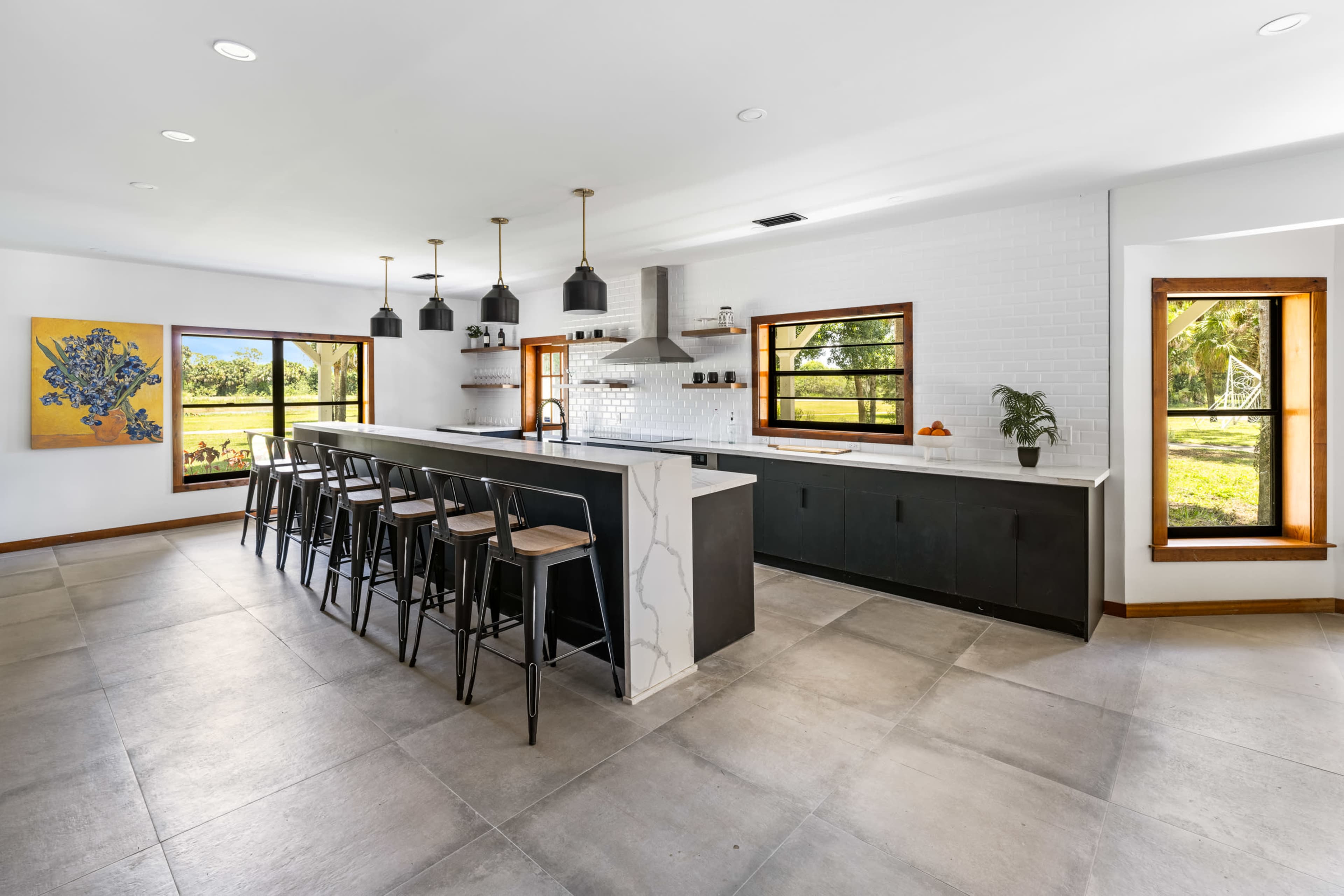 A modern kitchen with a large island, black bar stools, and windows showing a green landscape outside.