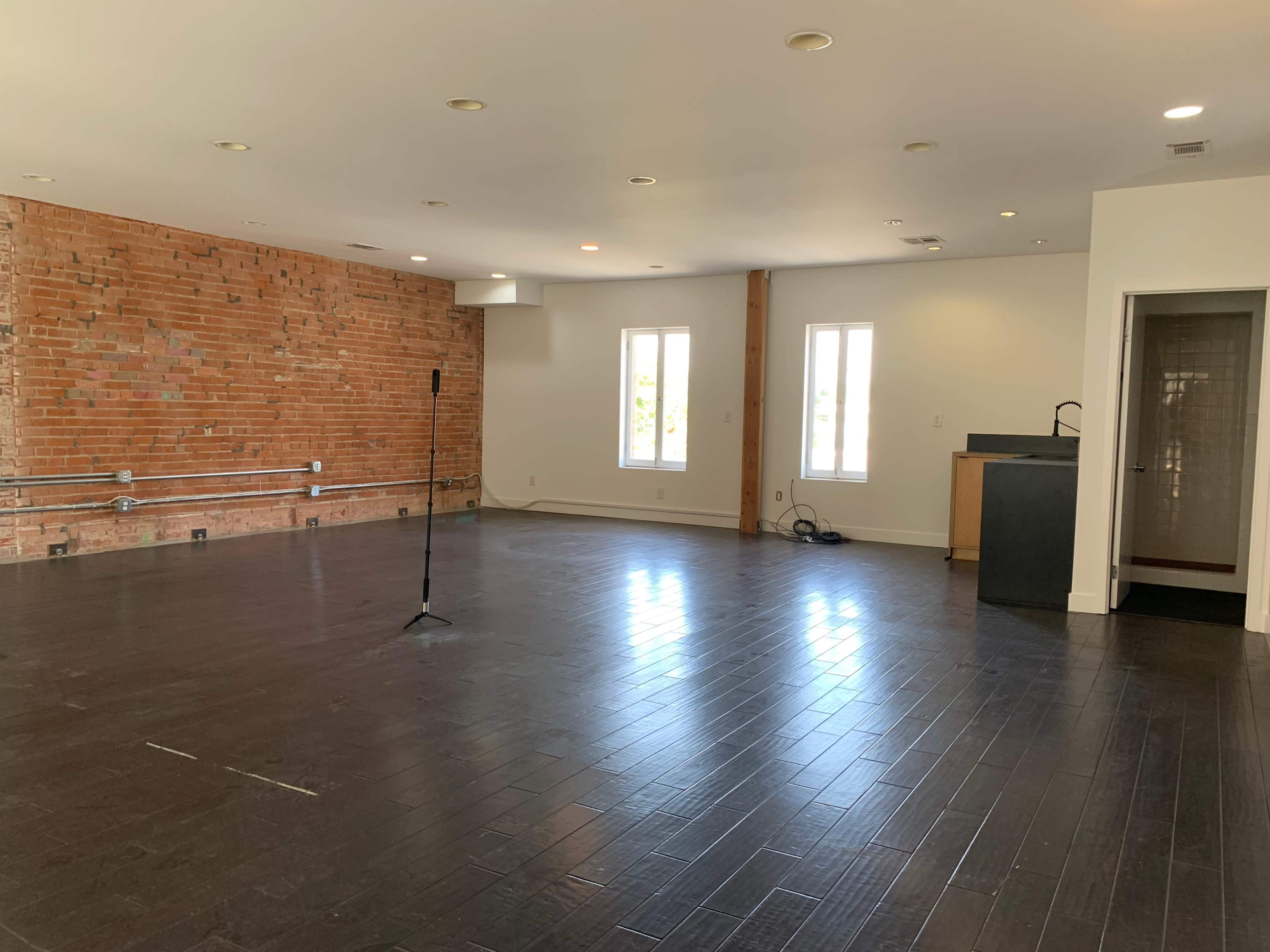 A spacious, open room with a brick accent wall, a microphone stand, and multiple windows allowing natural light.