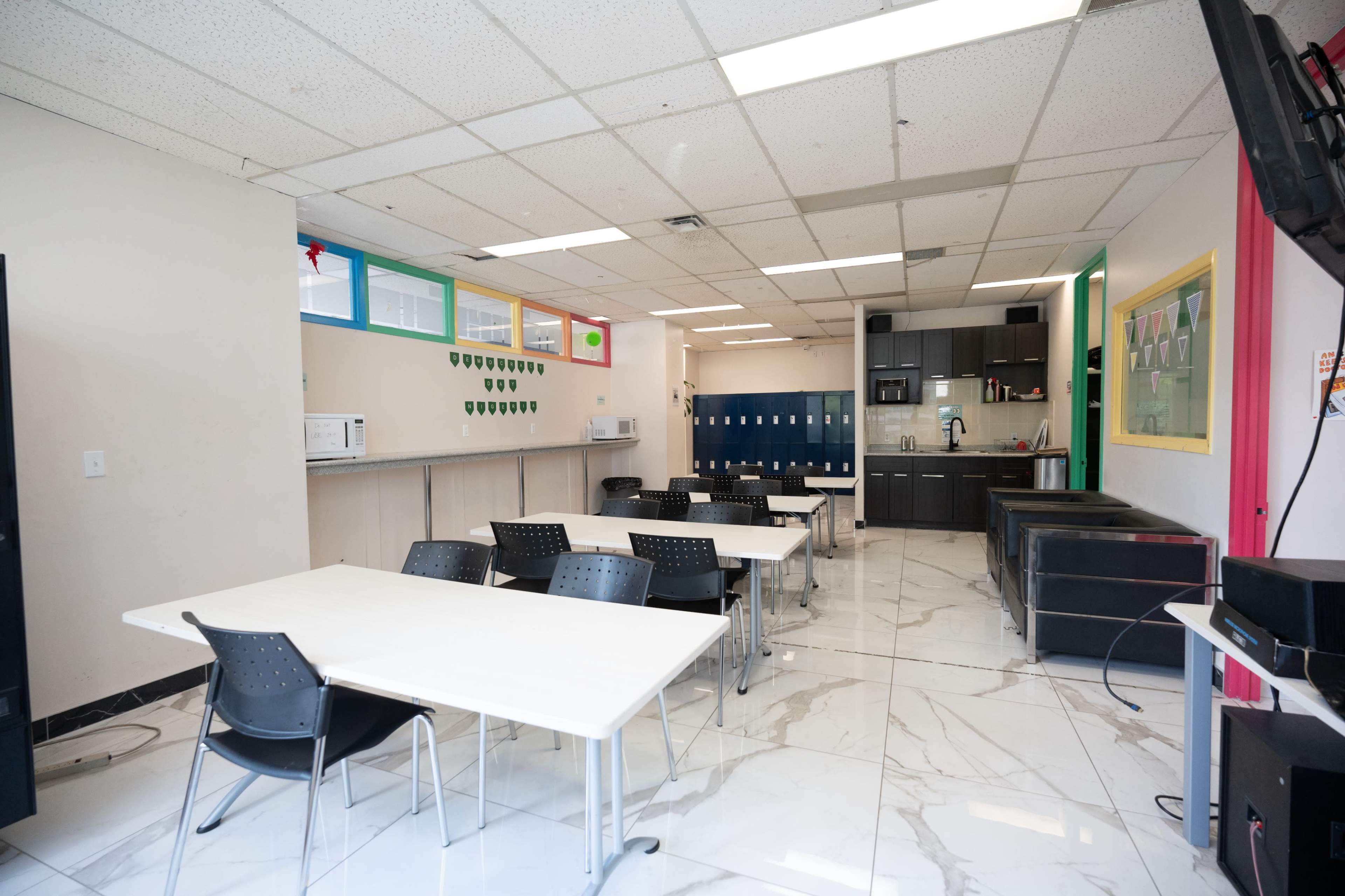 The image shows a spacious, well-lit classroom or break room with several white tables and black chairs, along with a kitchenette area and lockers against the wall.