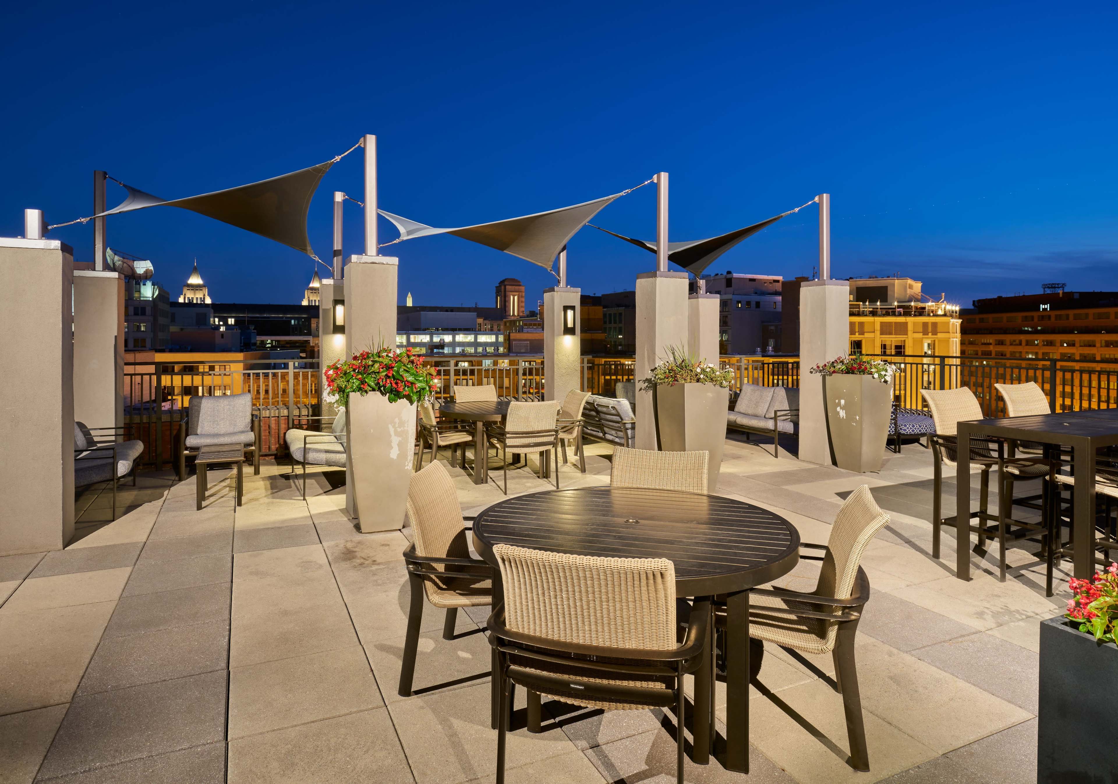 The rooftop terrace features several seating areas with tables and chairs, surrounded by planters and shaded by canopies, against a backdrop of a twilight sky and city buildings.