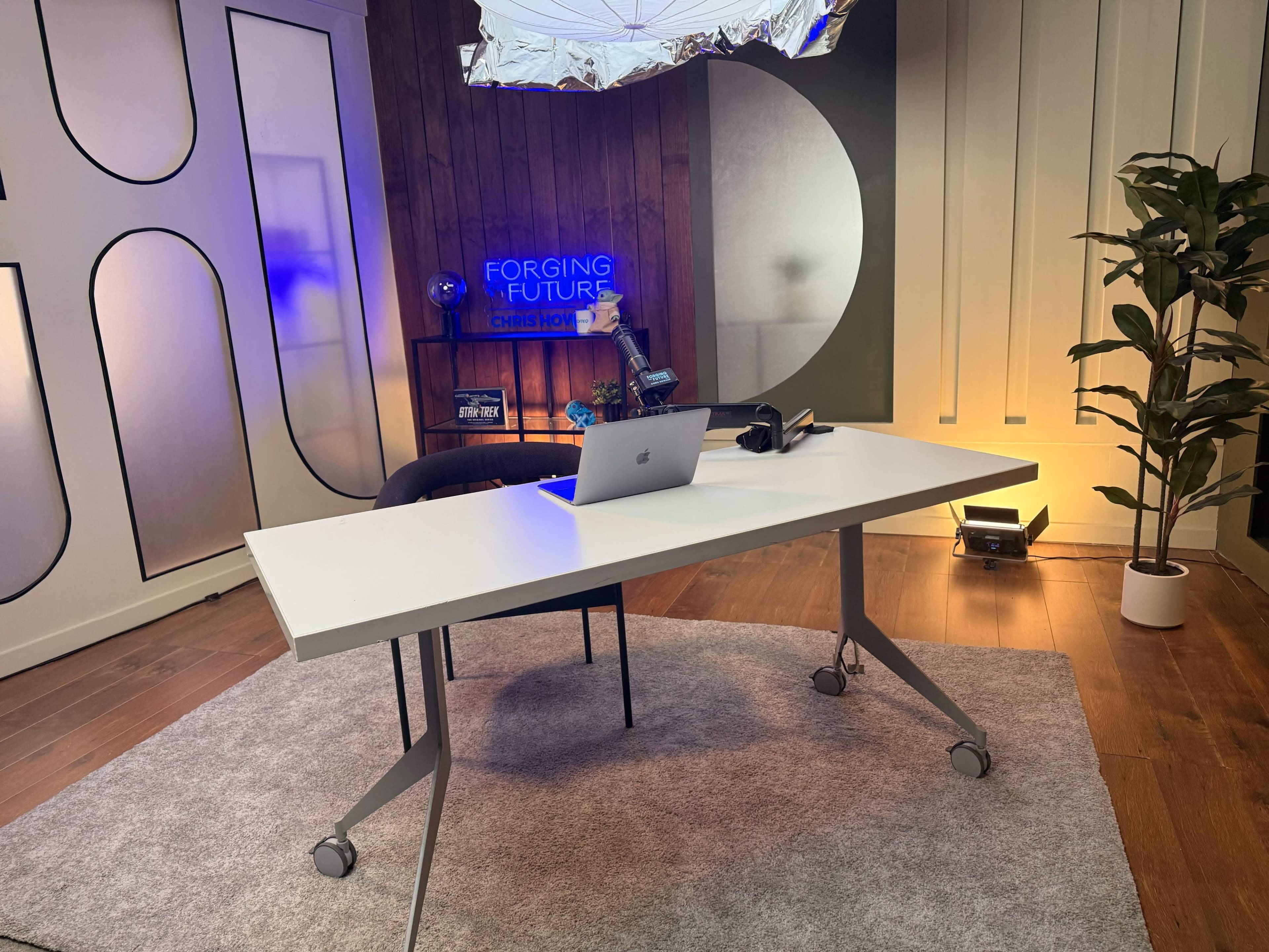 A modern, minimalist workspace features a white desk with a laptop, headphones, and a decorative plant, set against a backdrop with abstract designs and soft lighting.