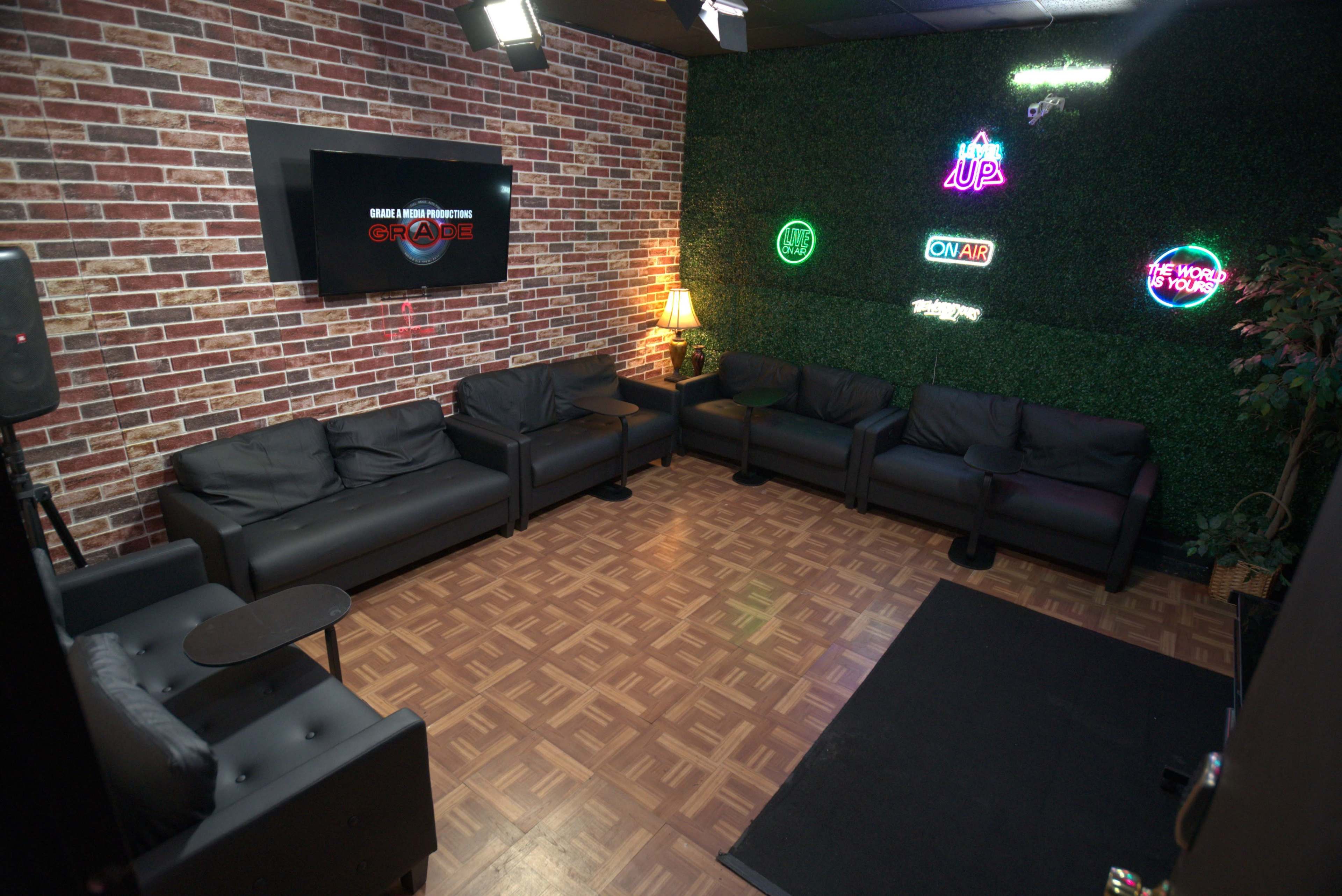 The image shows a small lounge area with black couches and a wooden floor, accented by a brick wall and green foliage, along with neon lights and a television screen.