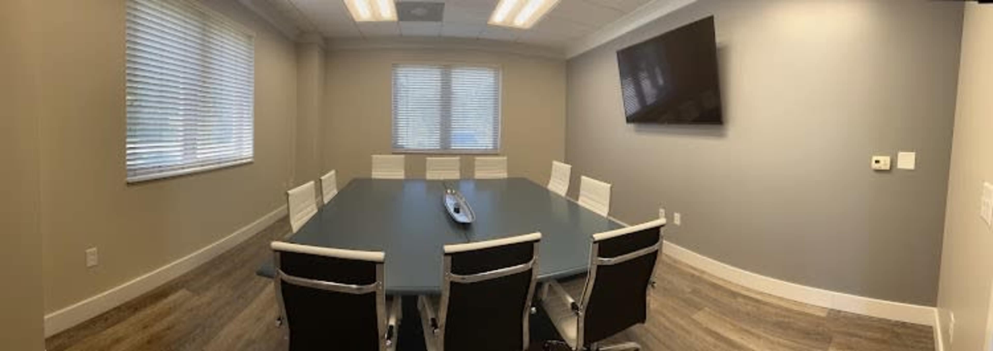 A modern conference room features a large table surrounded by white chairs, with a television mounted on the gray wall and windows allowing natural light.