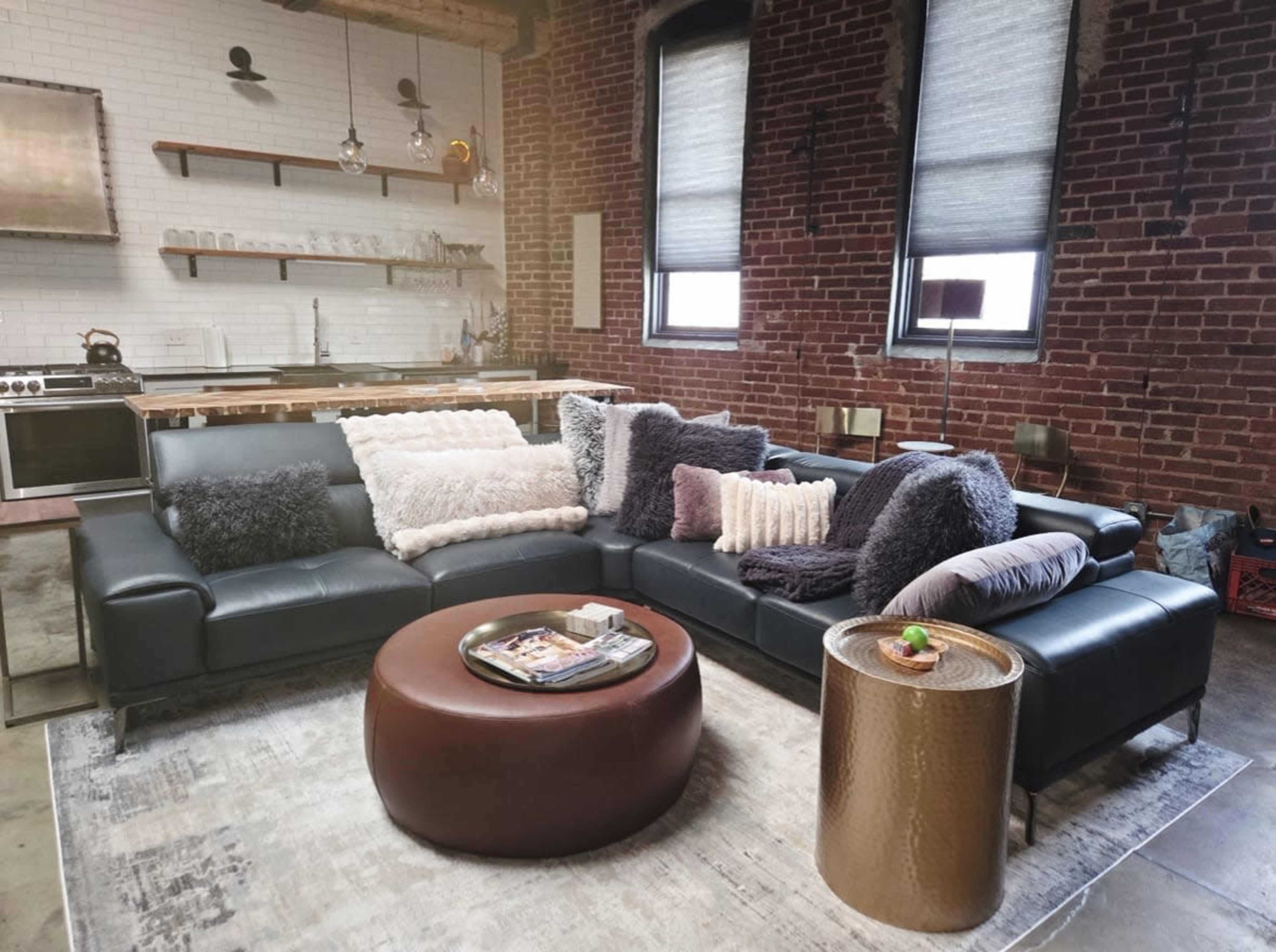 The image shows a stylish living room with a dark sectional sofa adorned with various textured pillows, a round coffee table, and a kitchen area in the background featuring exposed brick walls and modern appliances.