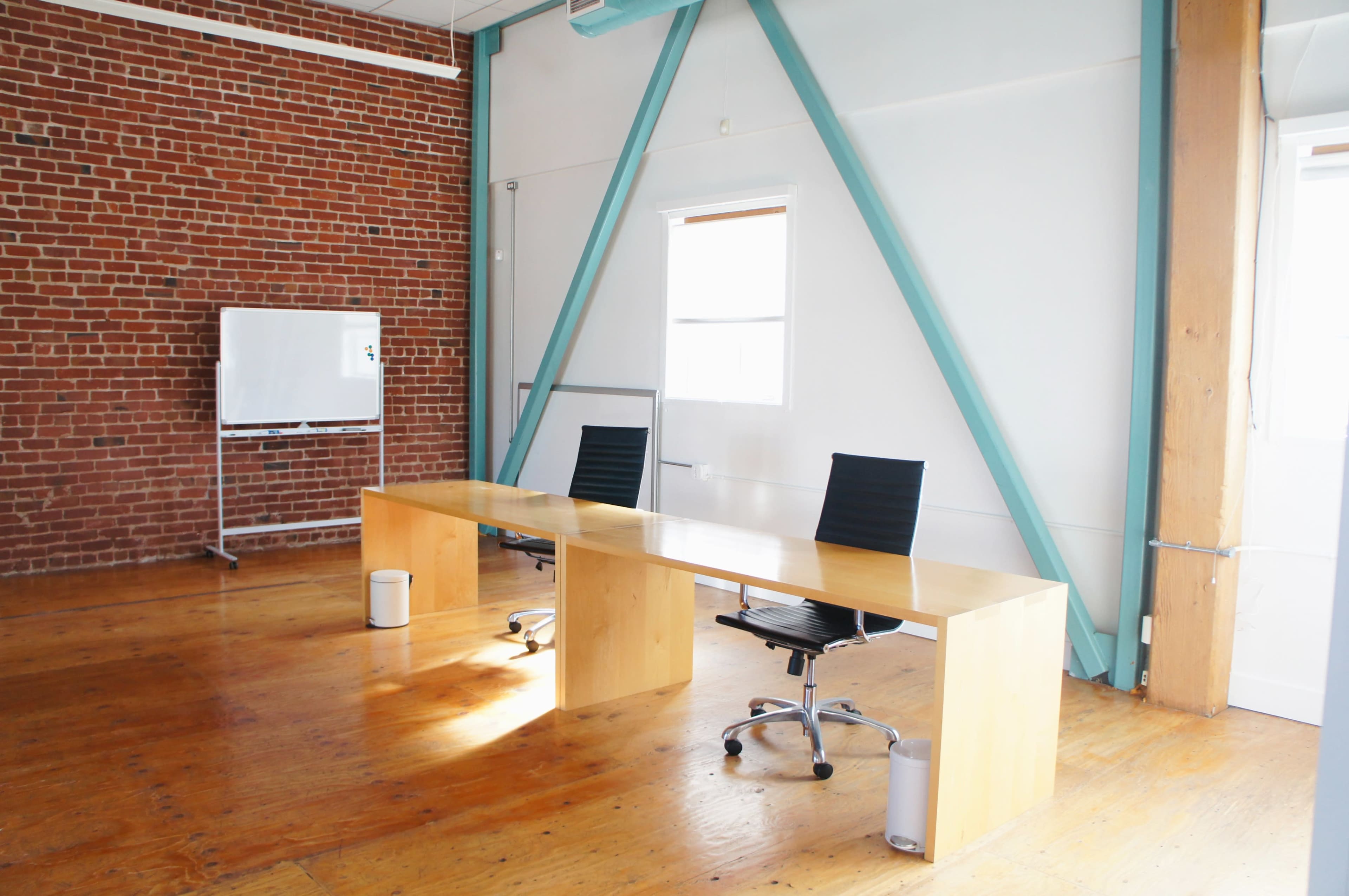 The image shows a minimalist office space with a wooden desk, two black office chairs, a whiteboard on wheels, and exposed brick walls.