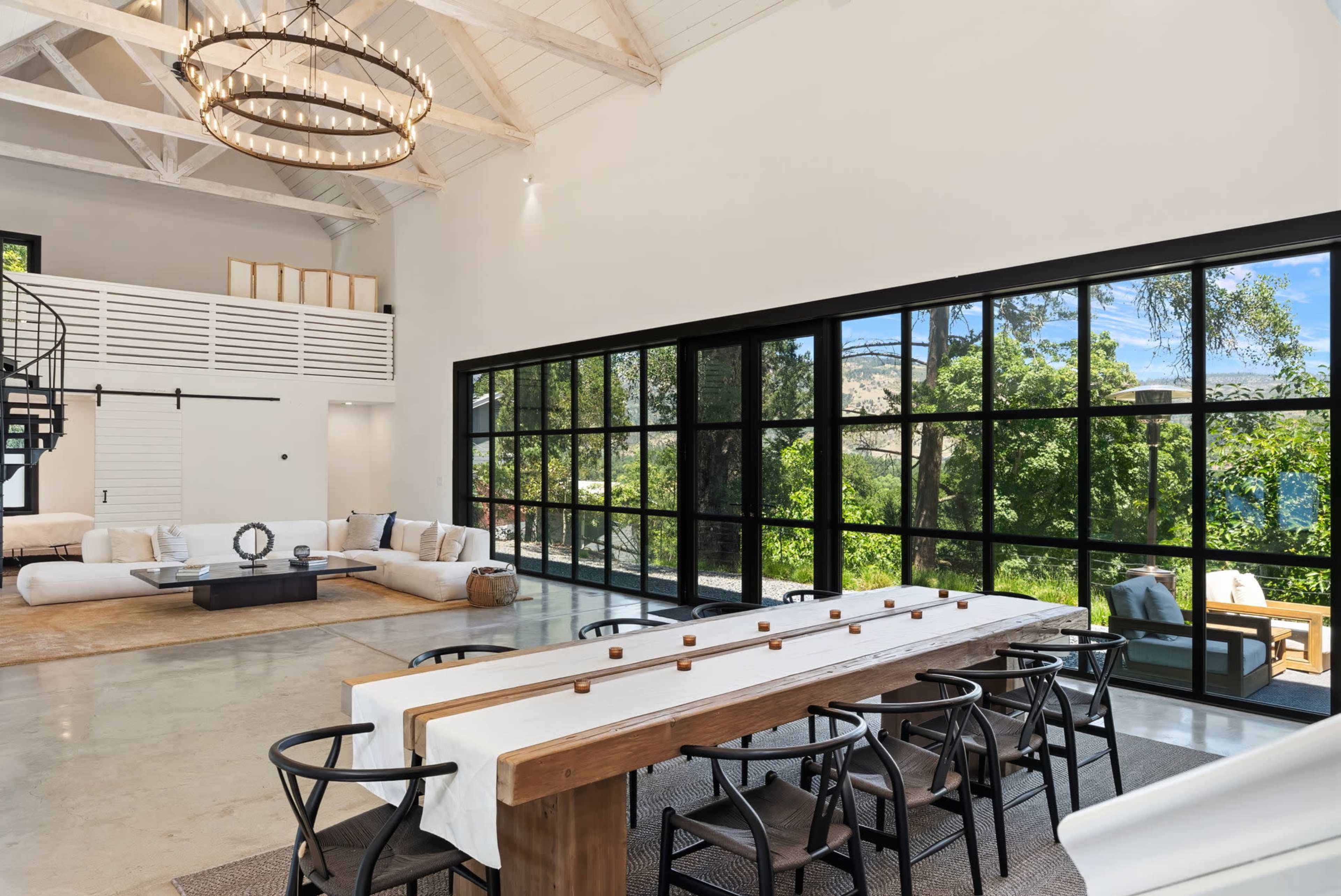 The image shows a spacious modern interior with a large dining table, a seating area, and floor-to-ceiling windows that provide a view of trees outside.
