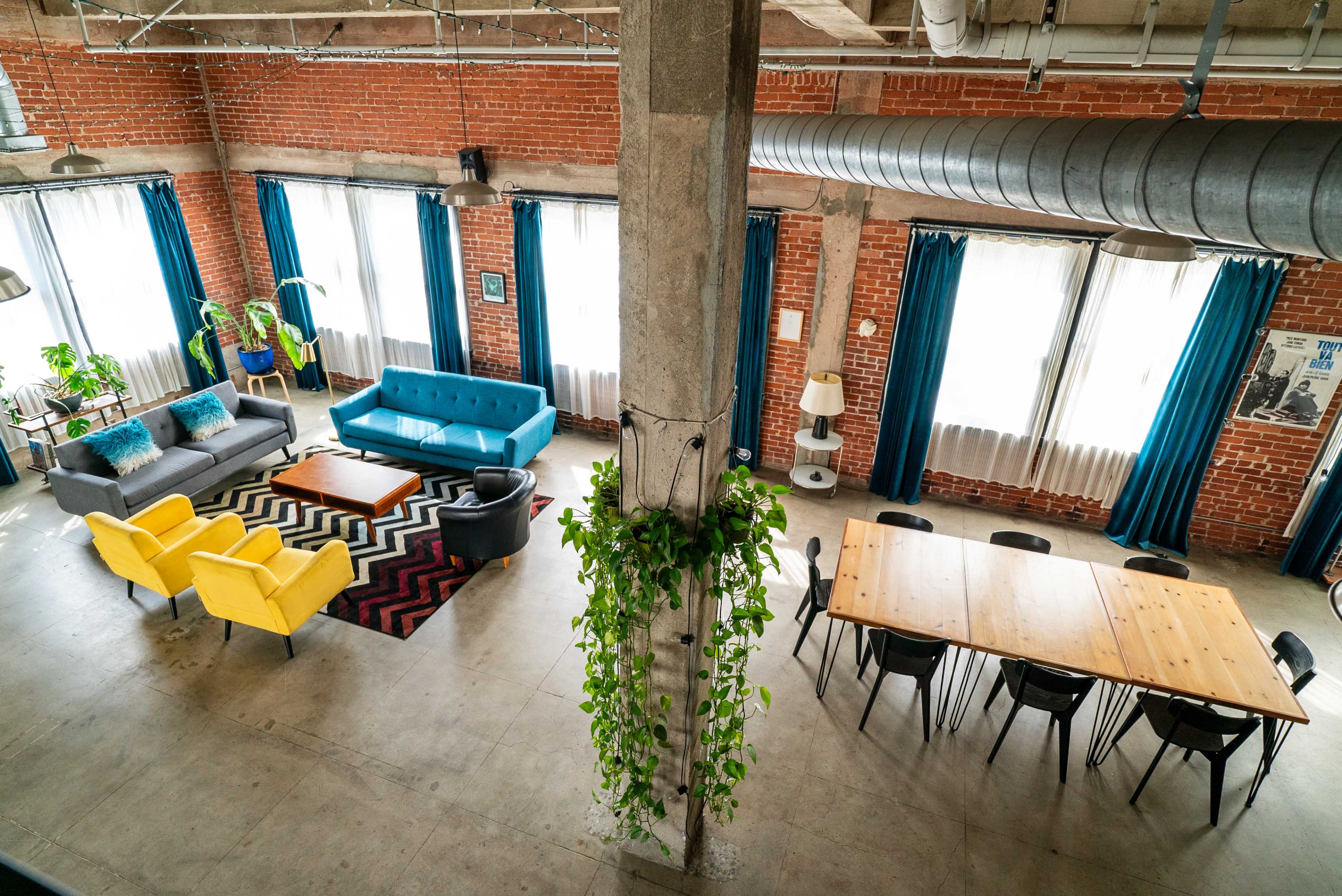 The image shows a spacious, modern interior with two seating areas furnished with colorful couches, a patterned rug, and a long wooden dining table surrounded by black chairs.