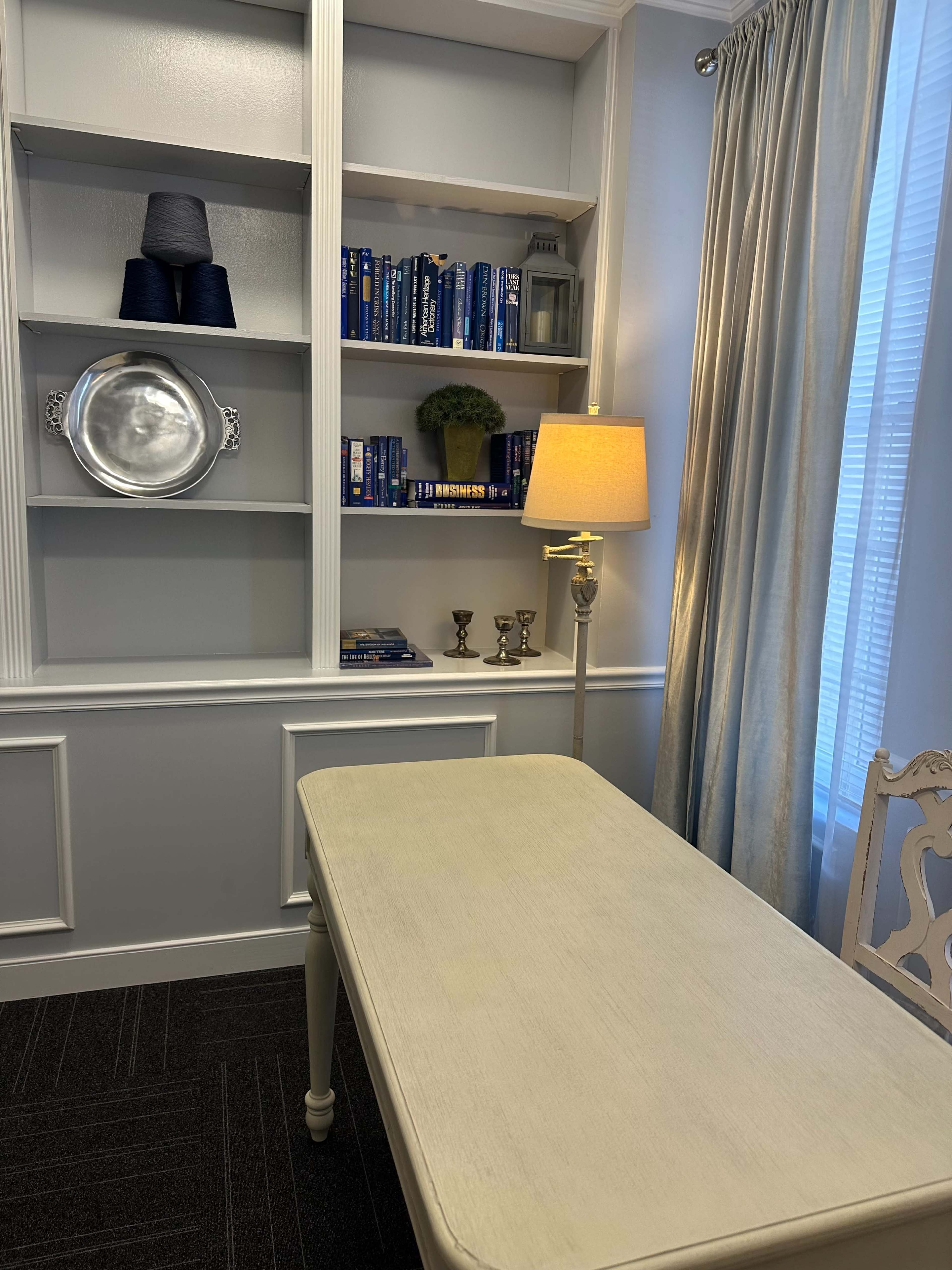 The image shows a small, well-organized room featuring a simple table in front of a bookshelf with books, a decorative plant, and a lamp.