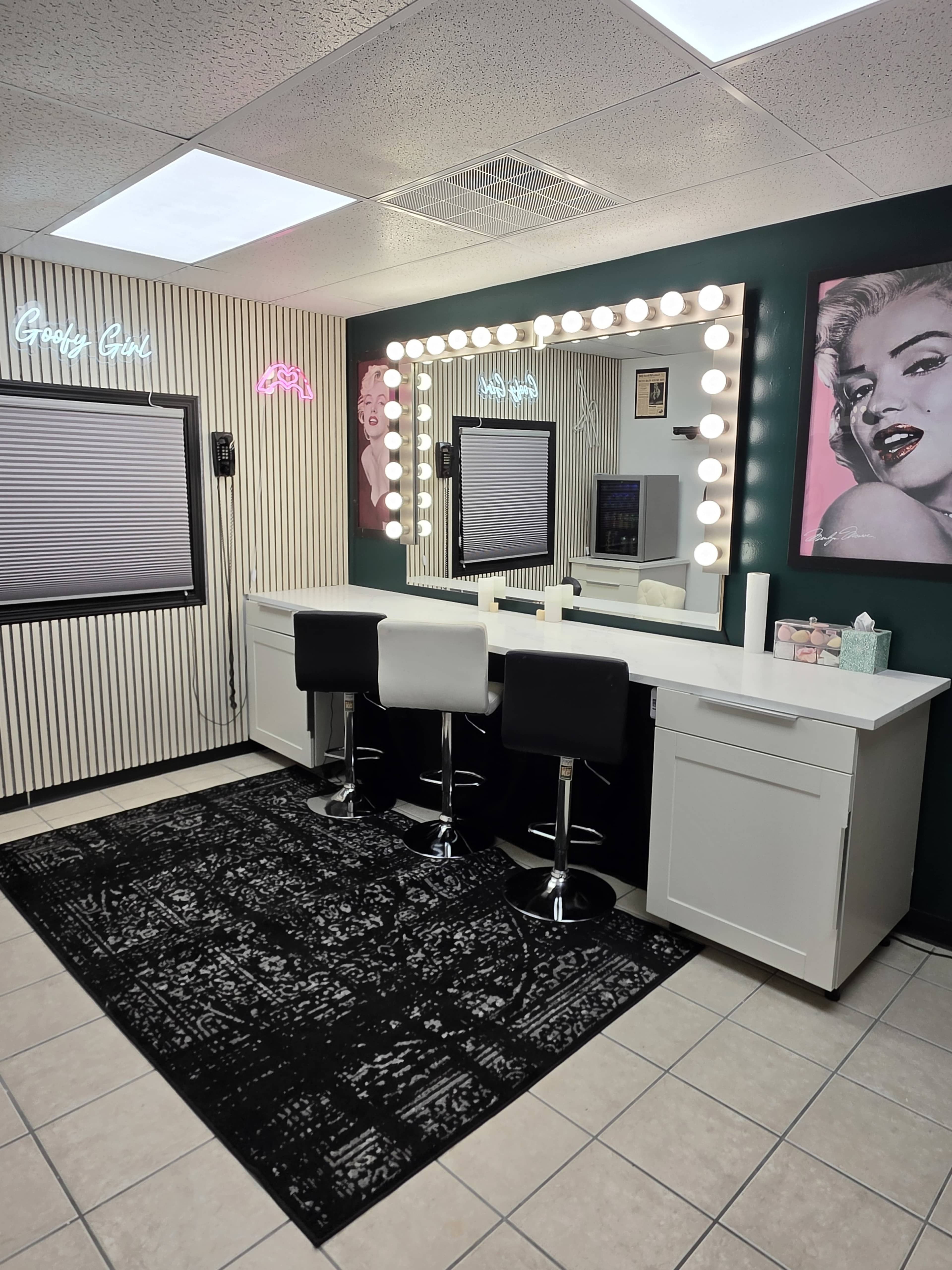 The image shows a stylish makeup vanity area with a large lighted mirror, black and white high stools, and a wall decorated with framed art and neon signs.