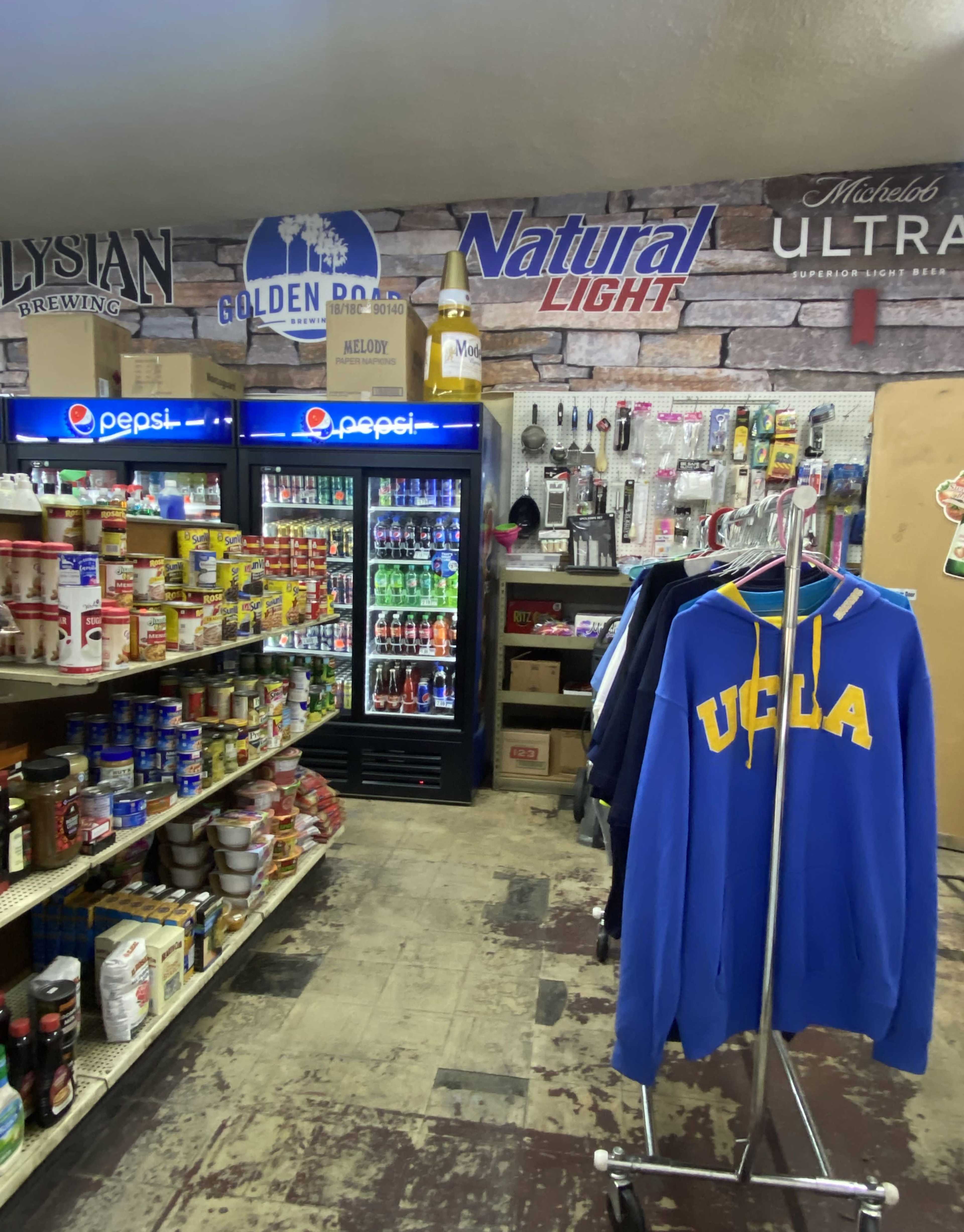 An aisle in a small store featuring shelves filled with various food products, a beverage cooler displaying drinks, and a rack of clothing, including a UCLA hoodie.