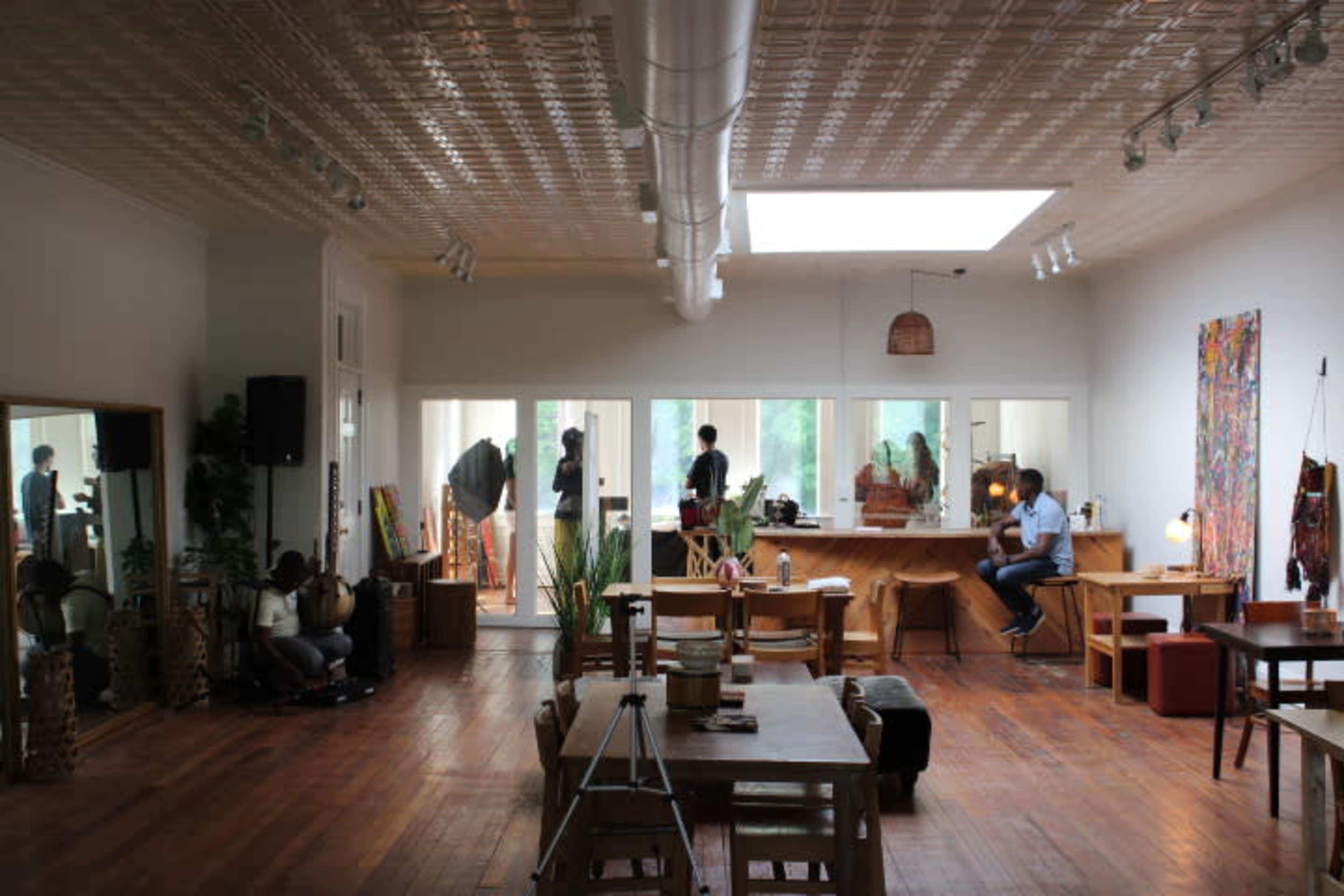 A spacious, well-lit room with wooden floors, a large table, and various furniture pieces, including a couch and art displays.