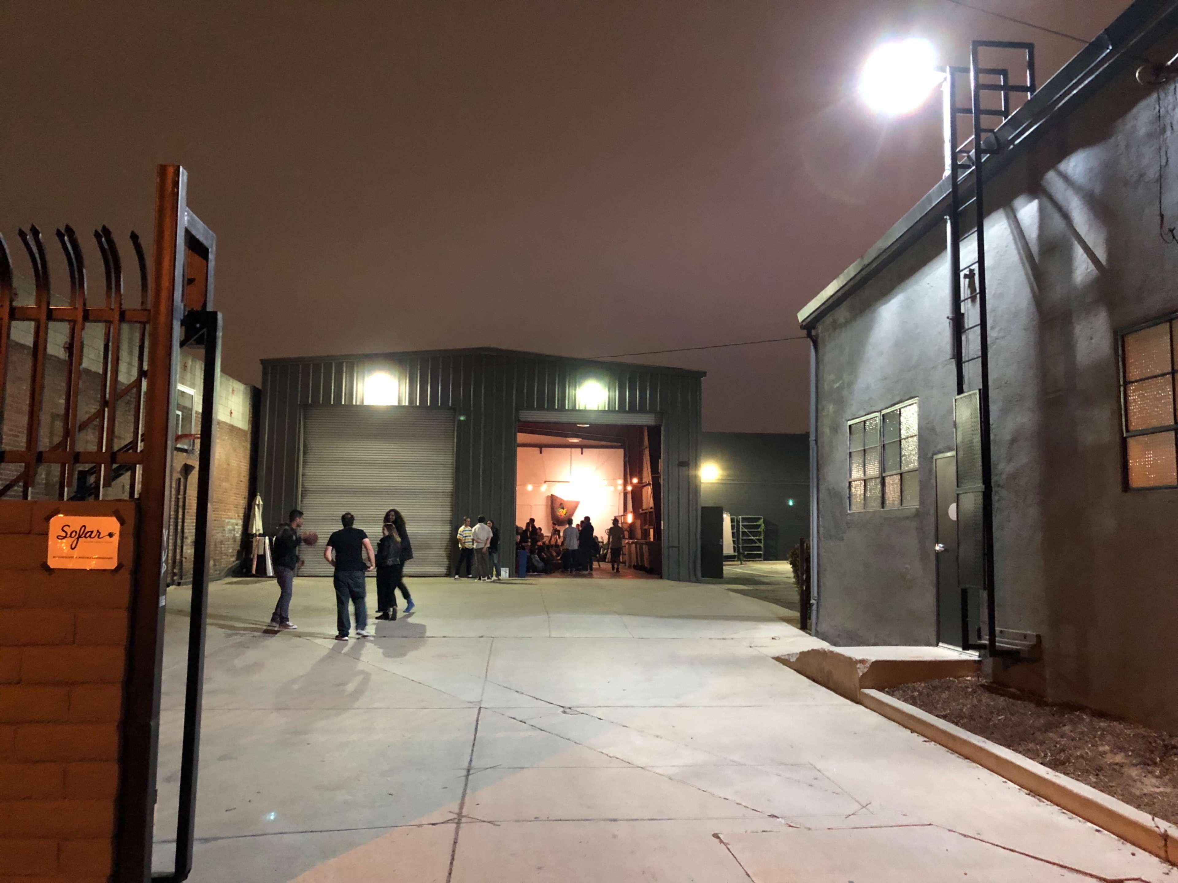 A group of people gathers outside a large metal warehouse entrance under dim lighting.