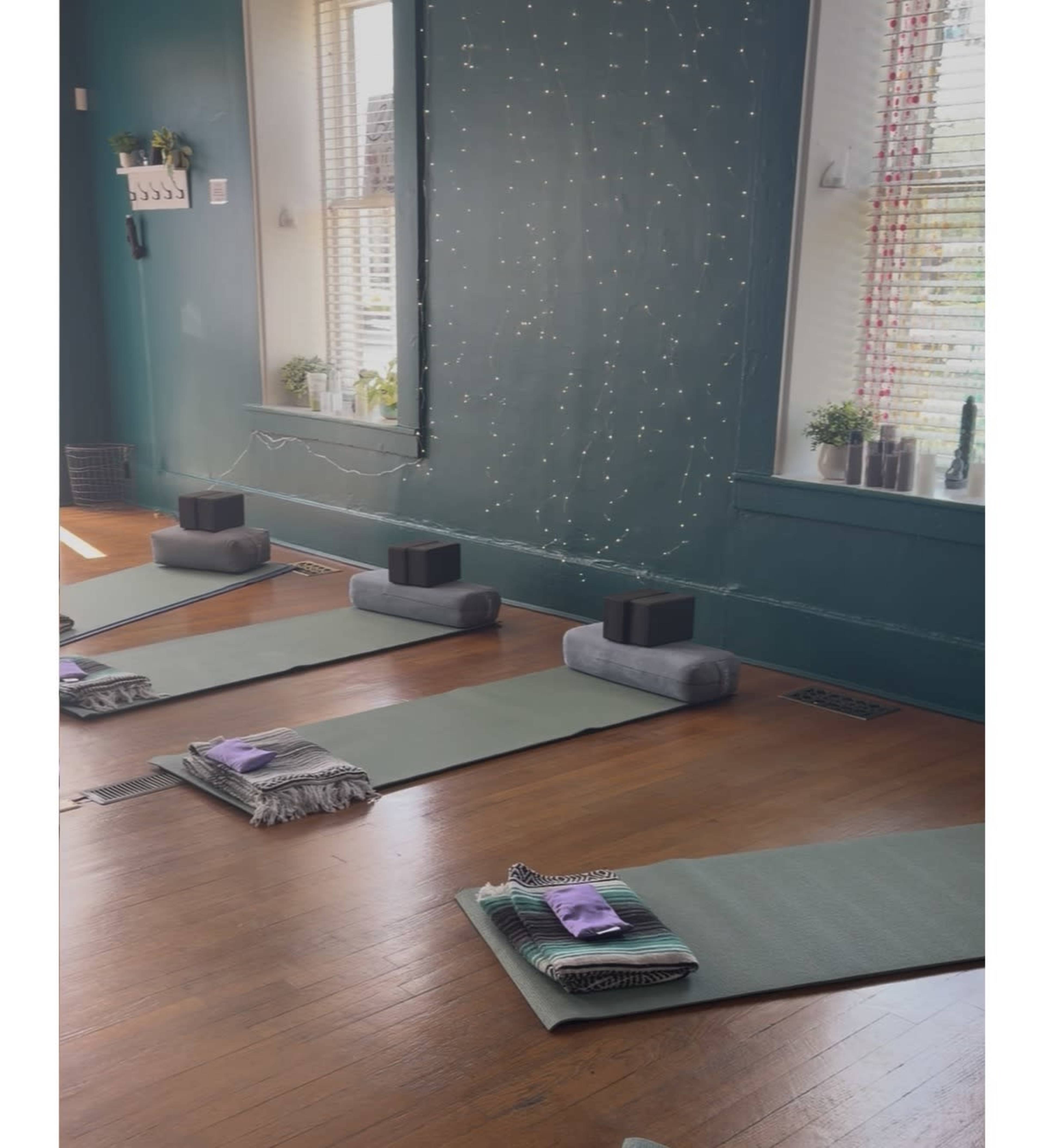 The image shows a yoga studio with neatly arranged mats, blocks, and rolled blankets placed on a wooden floor, accompanied by twinkling lights on the walls.