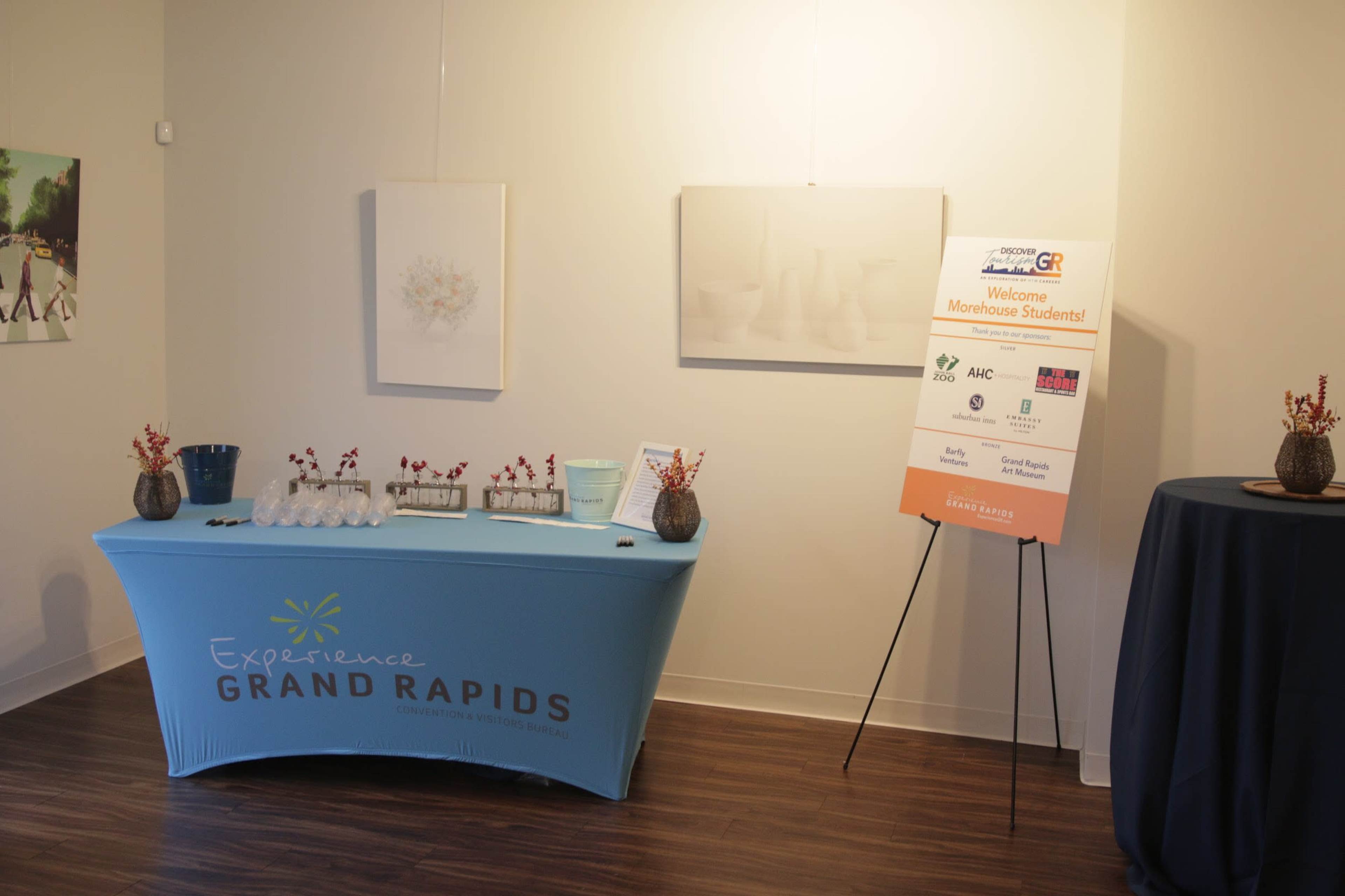 A table with a blue cover displaying "Experience Grand Rapids" is set up in a gallery, accompanied by a welcome sign for students and a backdrop of minimalistic artwork on the walls.