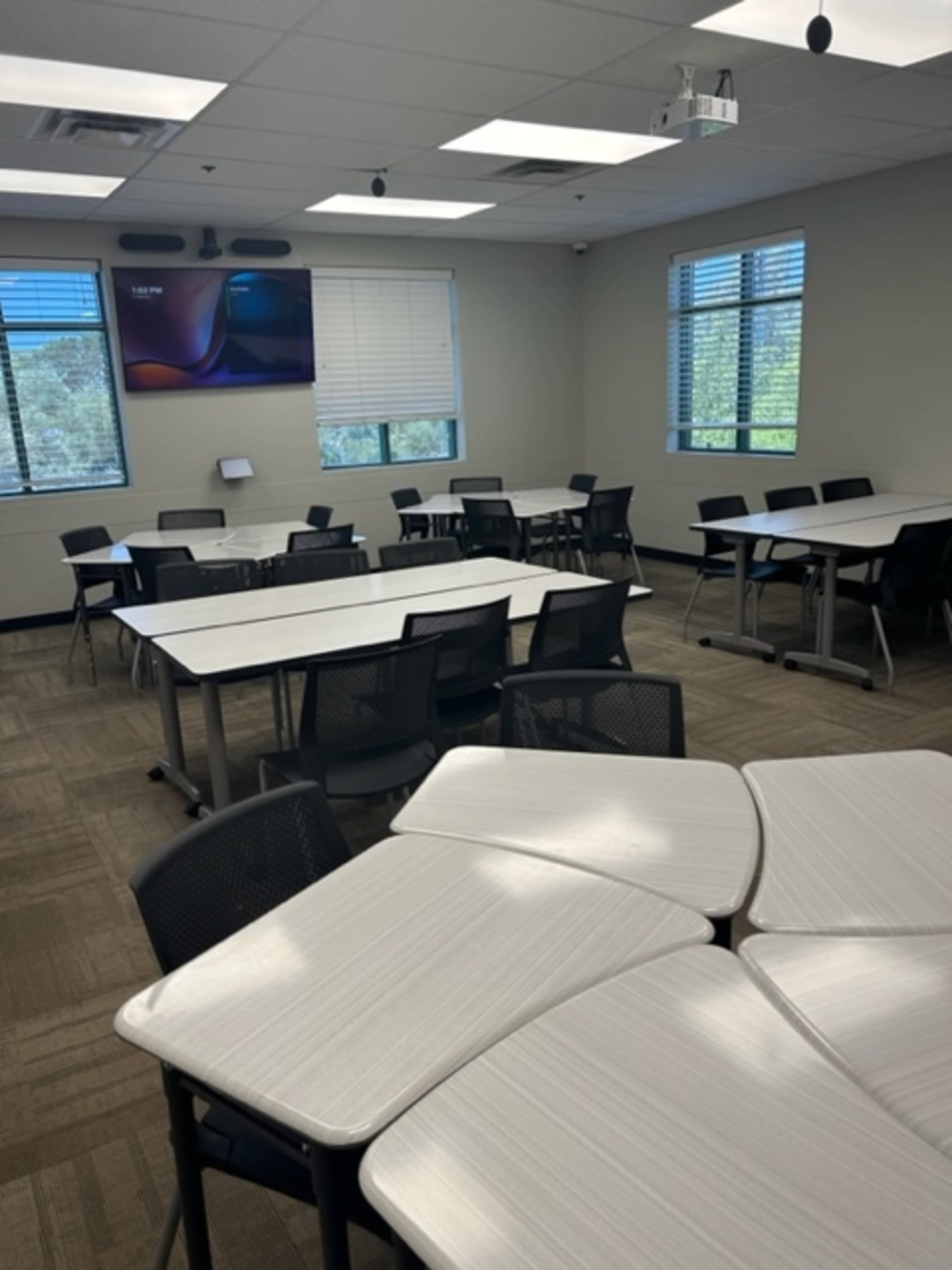 The image shows a modern classroom with multiple arranged desks and chairs, a mounted screen, and large windows allowing natural light.