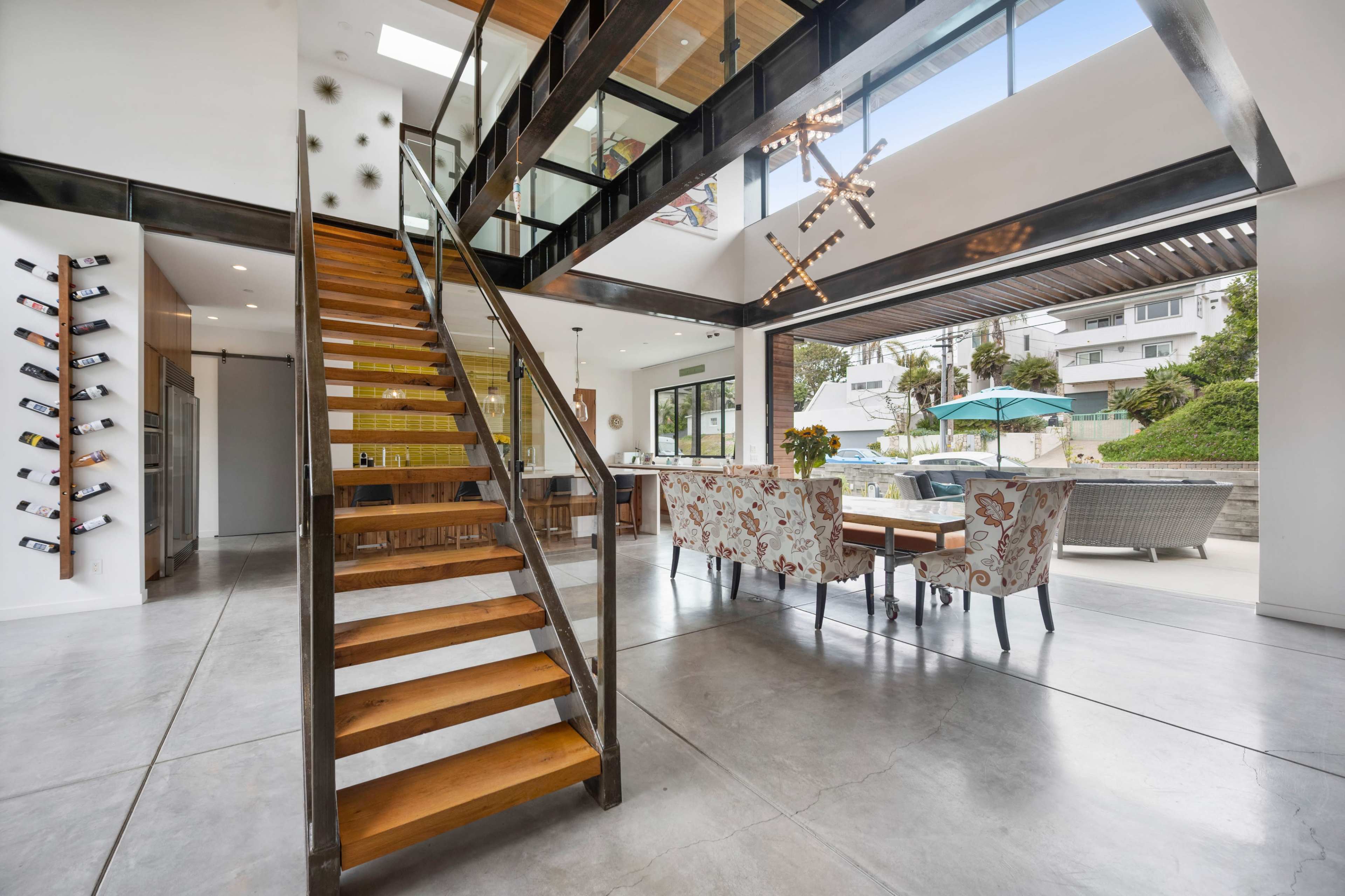 The image shows a modern interior space featuring a prominent wooden staircase, an open dining area with floral chairs, and large windows that provide natural light and a view of an outdoor patio.