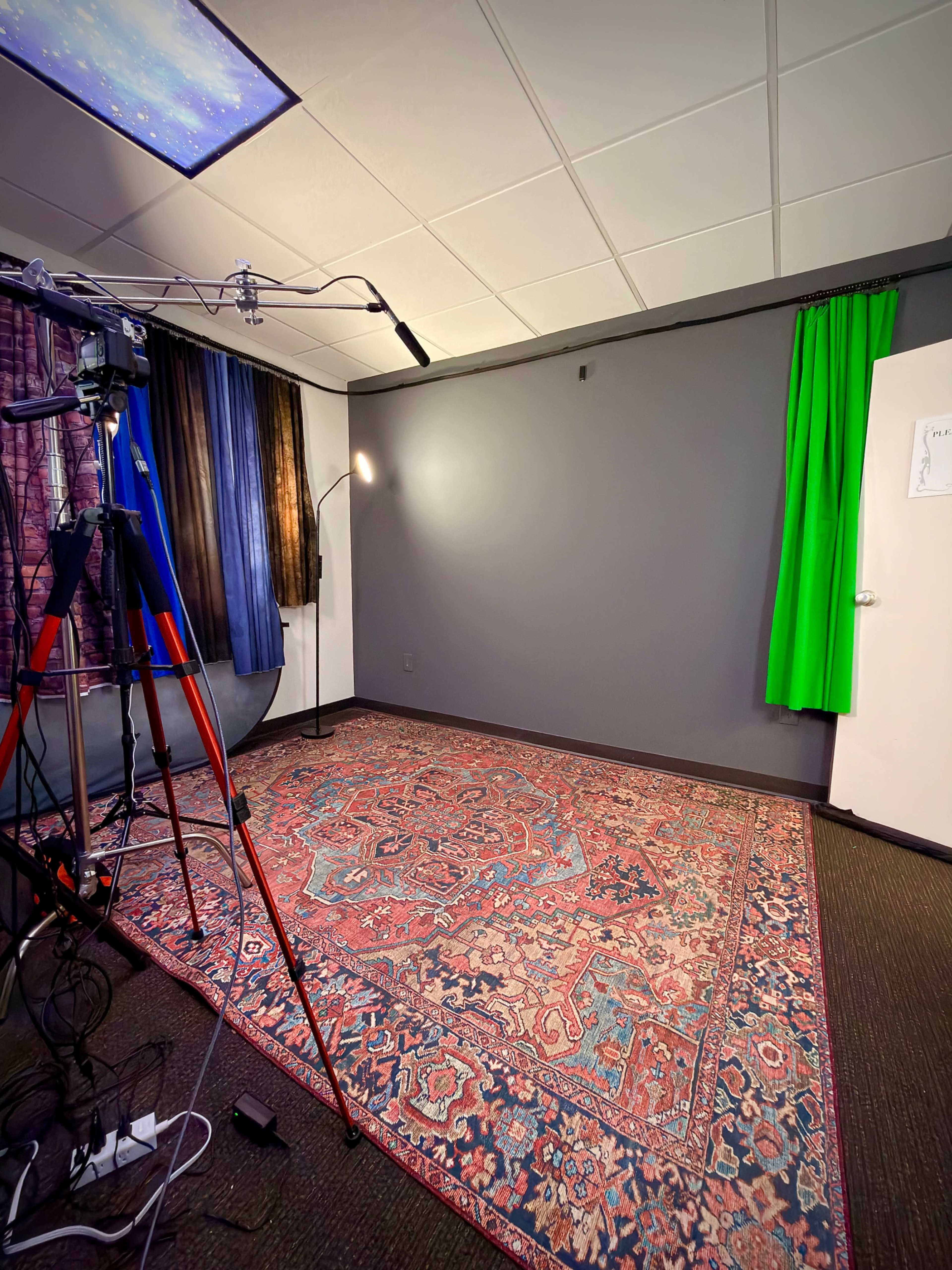 The image shows a spacious room with a patterned rug, a camera on a tripod, a standing lamp, two sets of curtains, and a green screen.