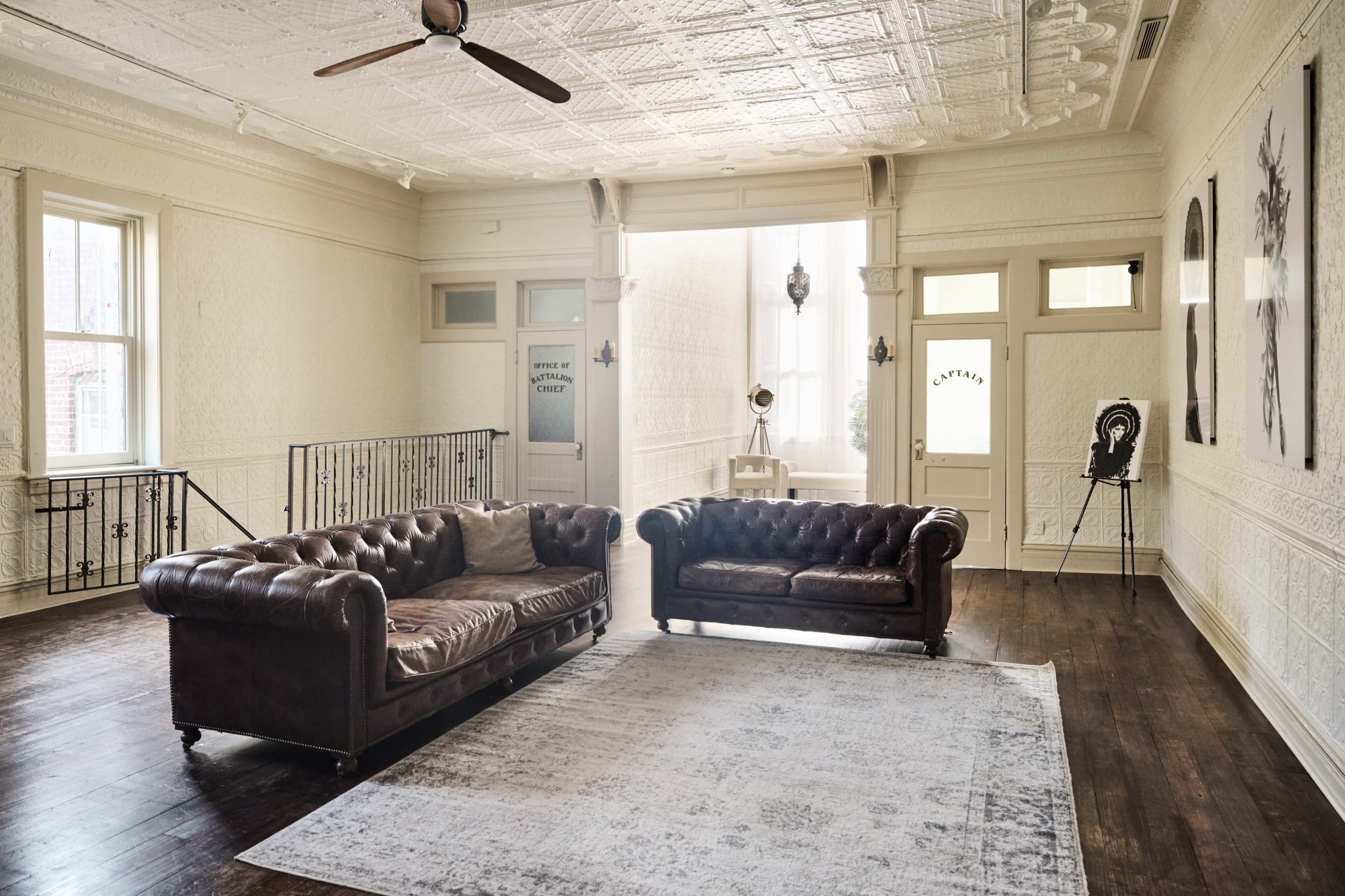 The room features two brown leather sofas and a light-colored area rug on a hardwood floor, with decorative walls and large windows allowing natural light to fill the space.