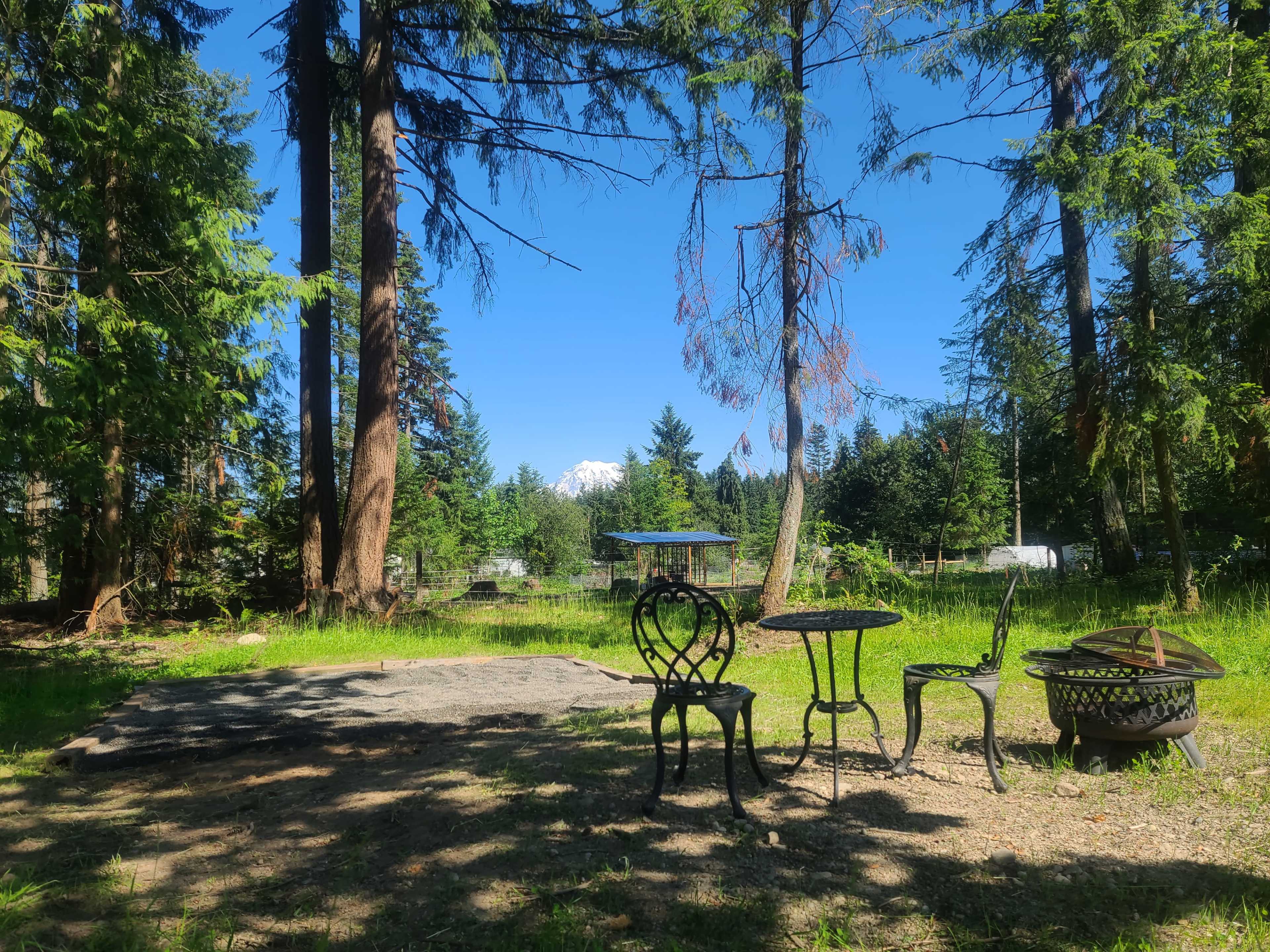 A rustic outdoor seating area with a small table and chairs is surrounded by tall trees and partial views of a mountain in the background.