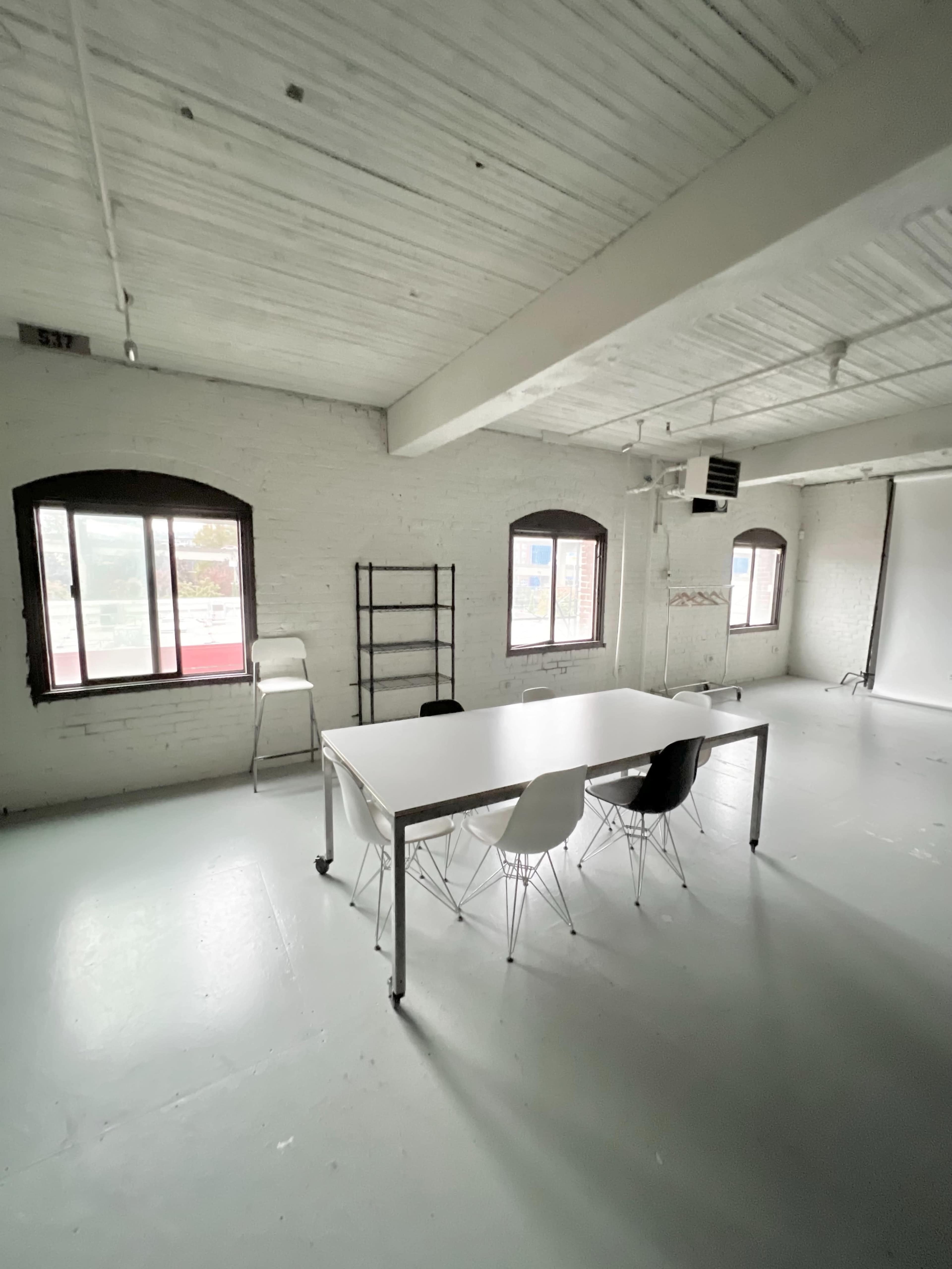 A sparse, well-lit room features a white table and four chairs set against textured walls with large windows.